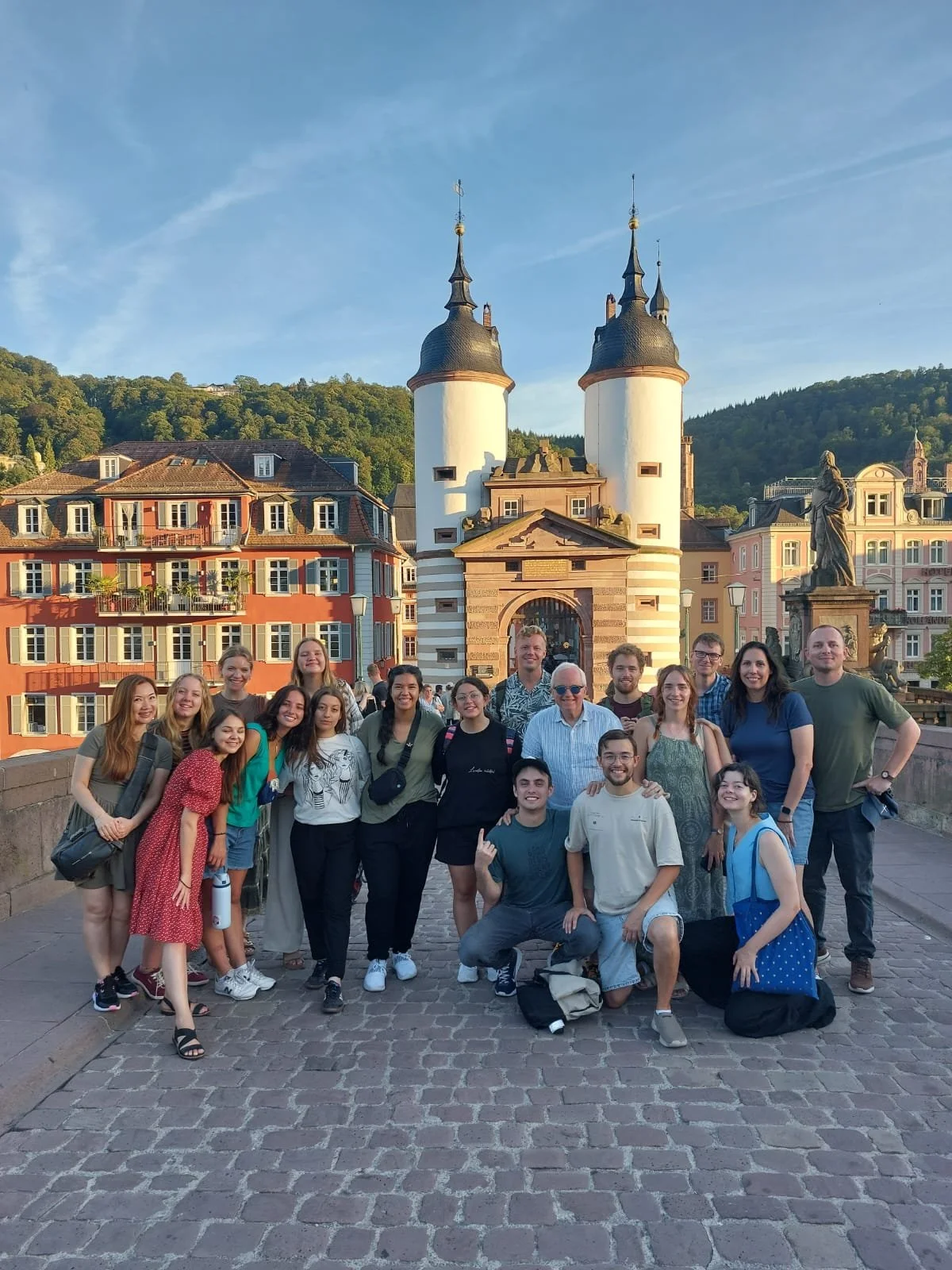 Group of diverse people smiling and posing for a photo on a cobblestone street with colorful European-style buildings, a historic gate with tower-like structures, and a hillside with trees in the background.