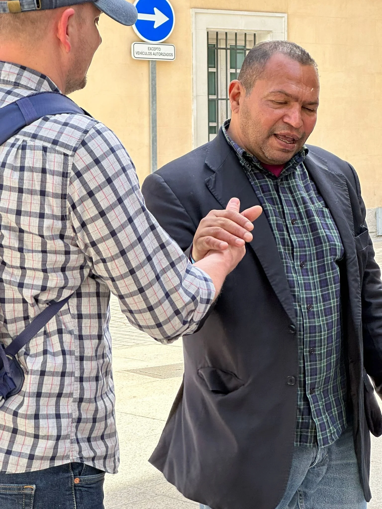Two men standing outdoors, engaged in conversation. One man is wearing a plaid shirt and a gray cap and has a backpack. The other man is wearing a dark jacket and a plaid shirt. There are street signs behind them, one of which indicates a one-way street in Spanish.