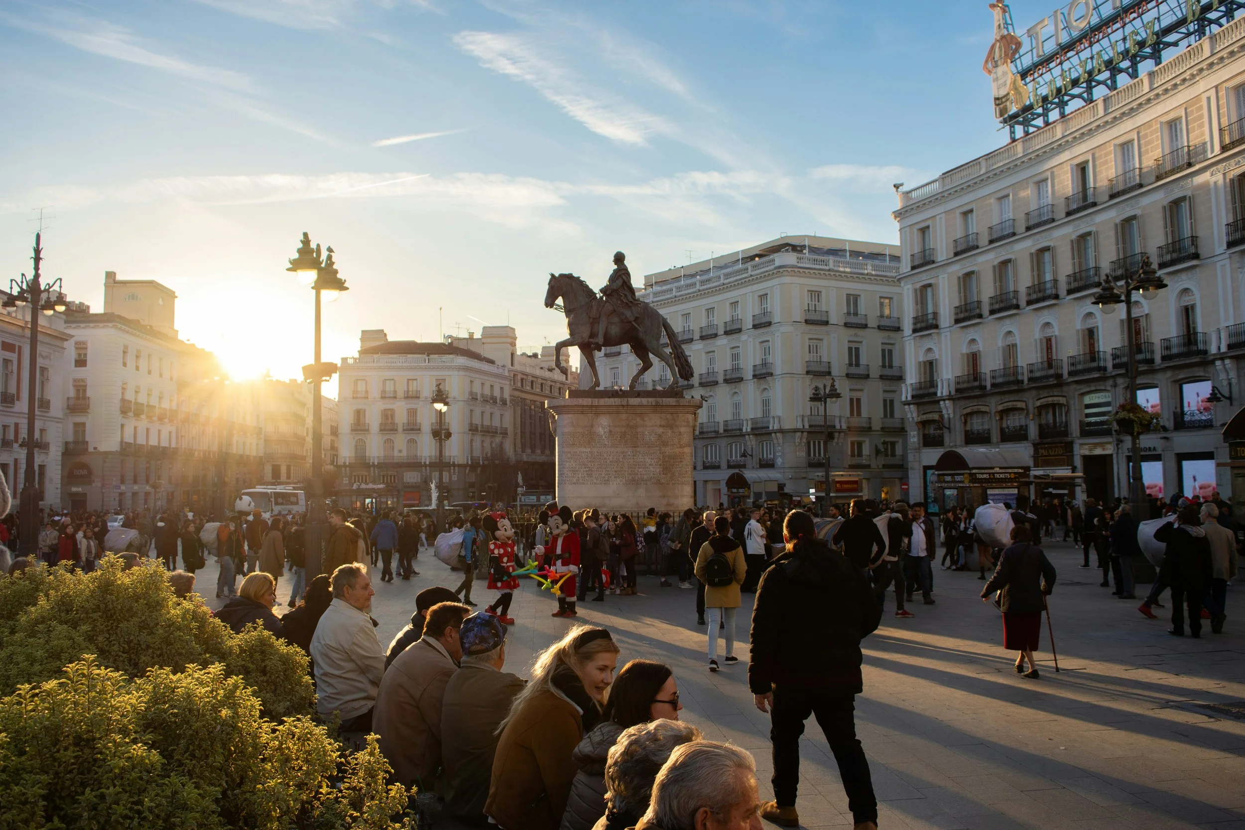 Sunset over a busy city square with a large equestrian statue at its center. Crowds of people, including children and adults, are gathered in front of the statue. Some children are dressed as Minnie Mouse characters. Surrounding buildings have multiple floors with balconies and historic architectural details. There are street lamps and a sign at the top of one building, with the sky partially cloudy.