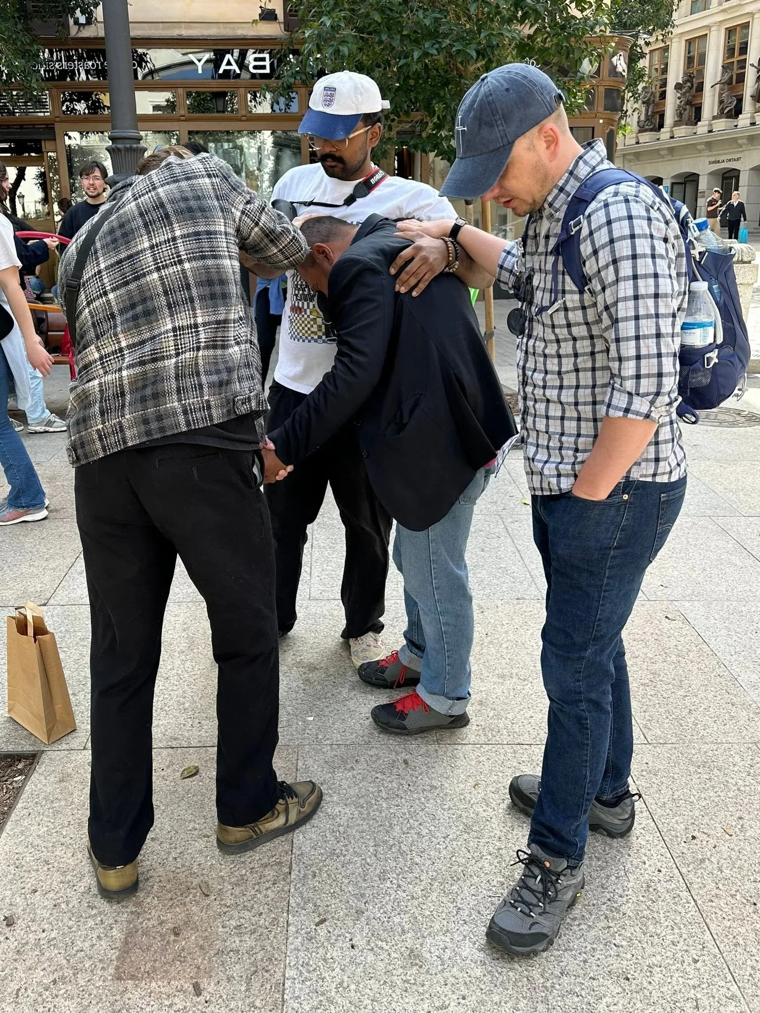 A group of people standing in a circle outdoors, holding hands in prayer or a moment of silence, with buildings and trees in the background.