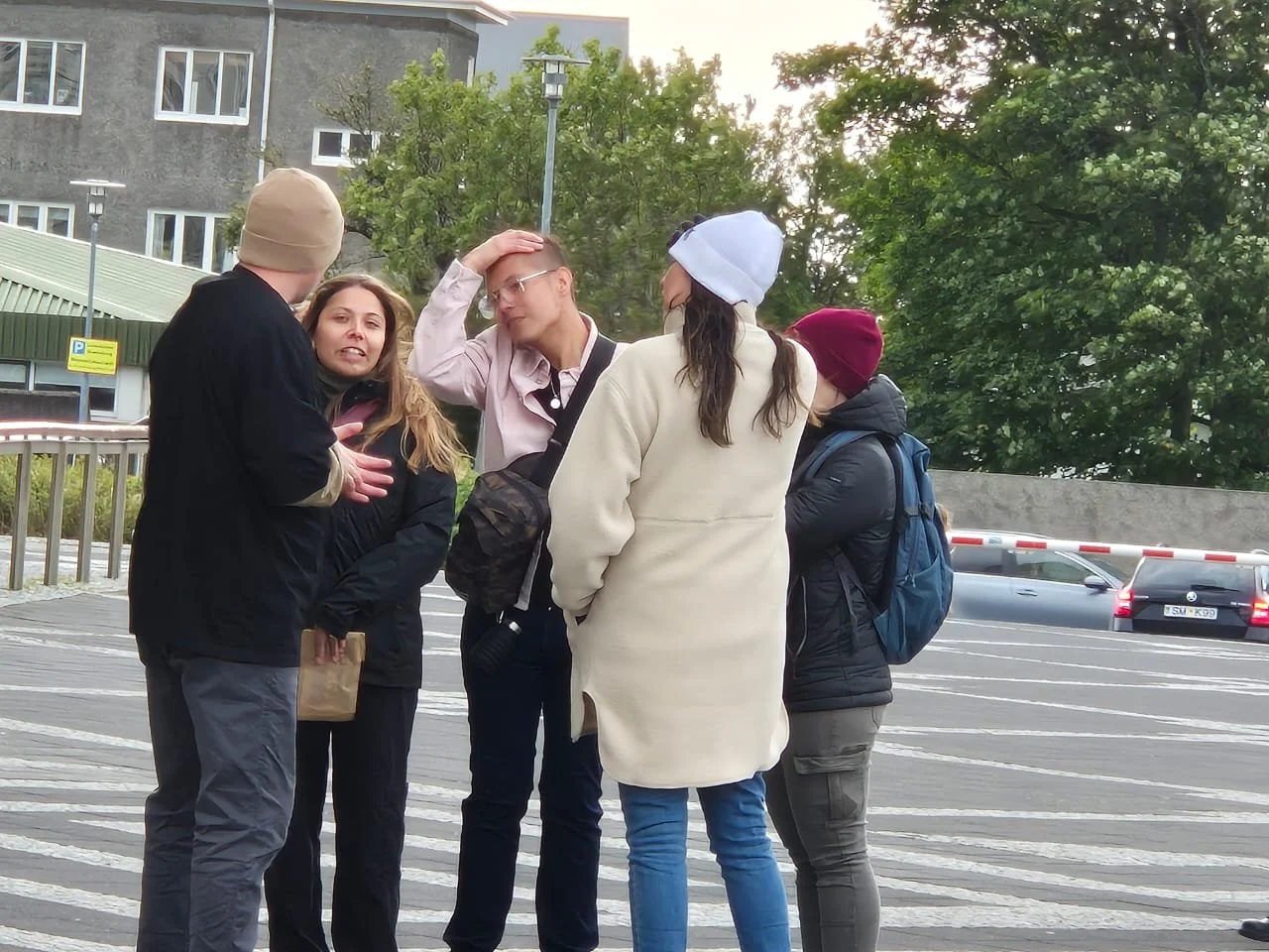 Group of five young adults having a conversation outdoors on a city street, with cars and trees in the background.