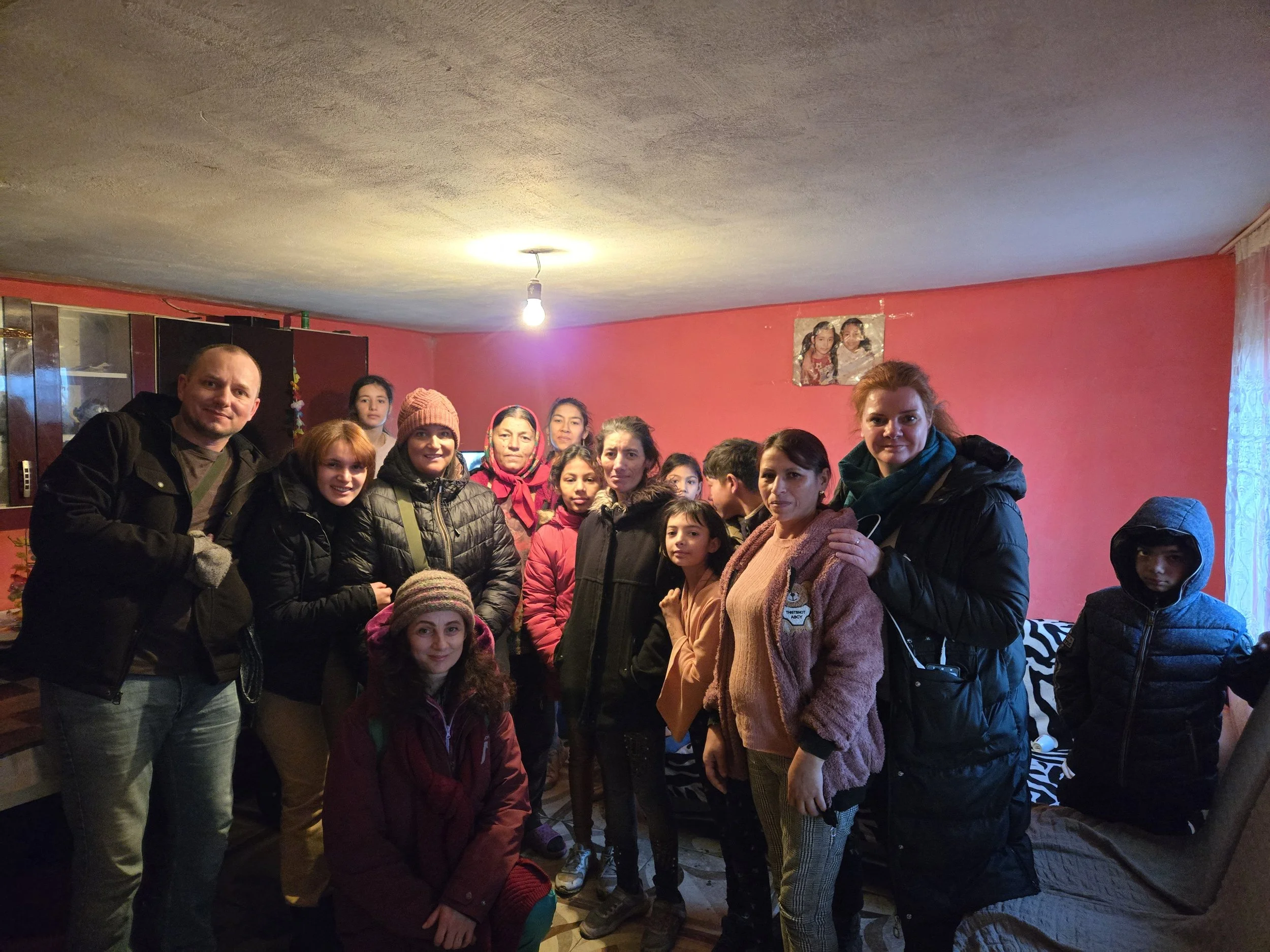 A group of people, including children and adults, gathered in a room with pink walls and a poster of children on the wall. Some are smiling and looking at the camera.