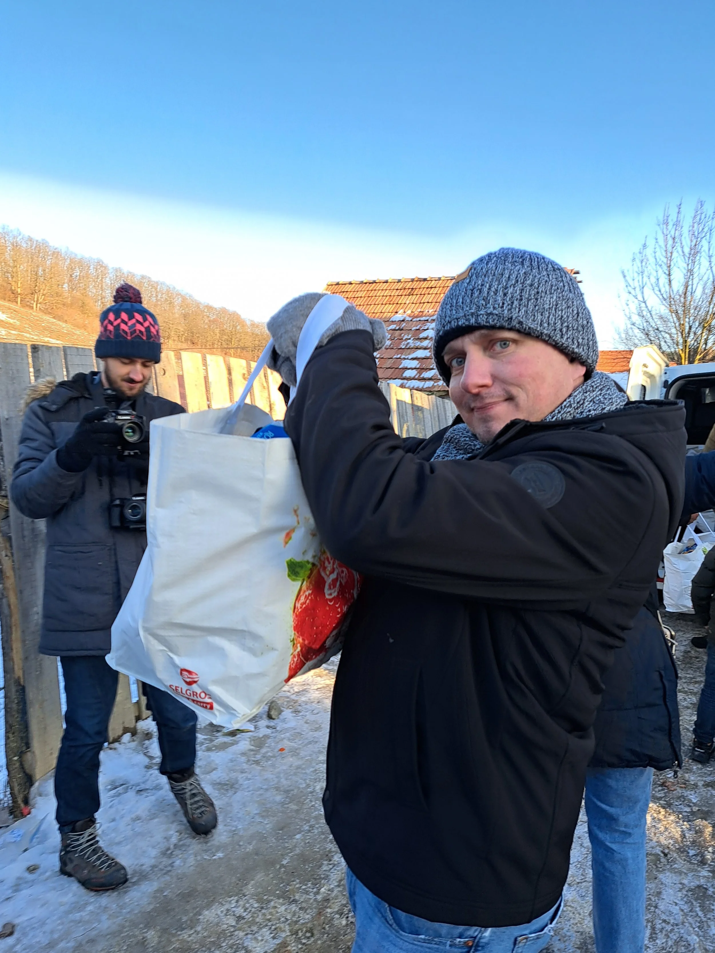 Group of men outside in winter, one smiling and handing a white shopping bag to another man. All are dressed warmly with hats and jackets, standing on snowy ground.