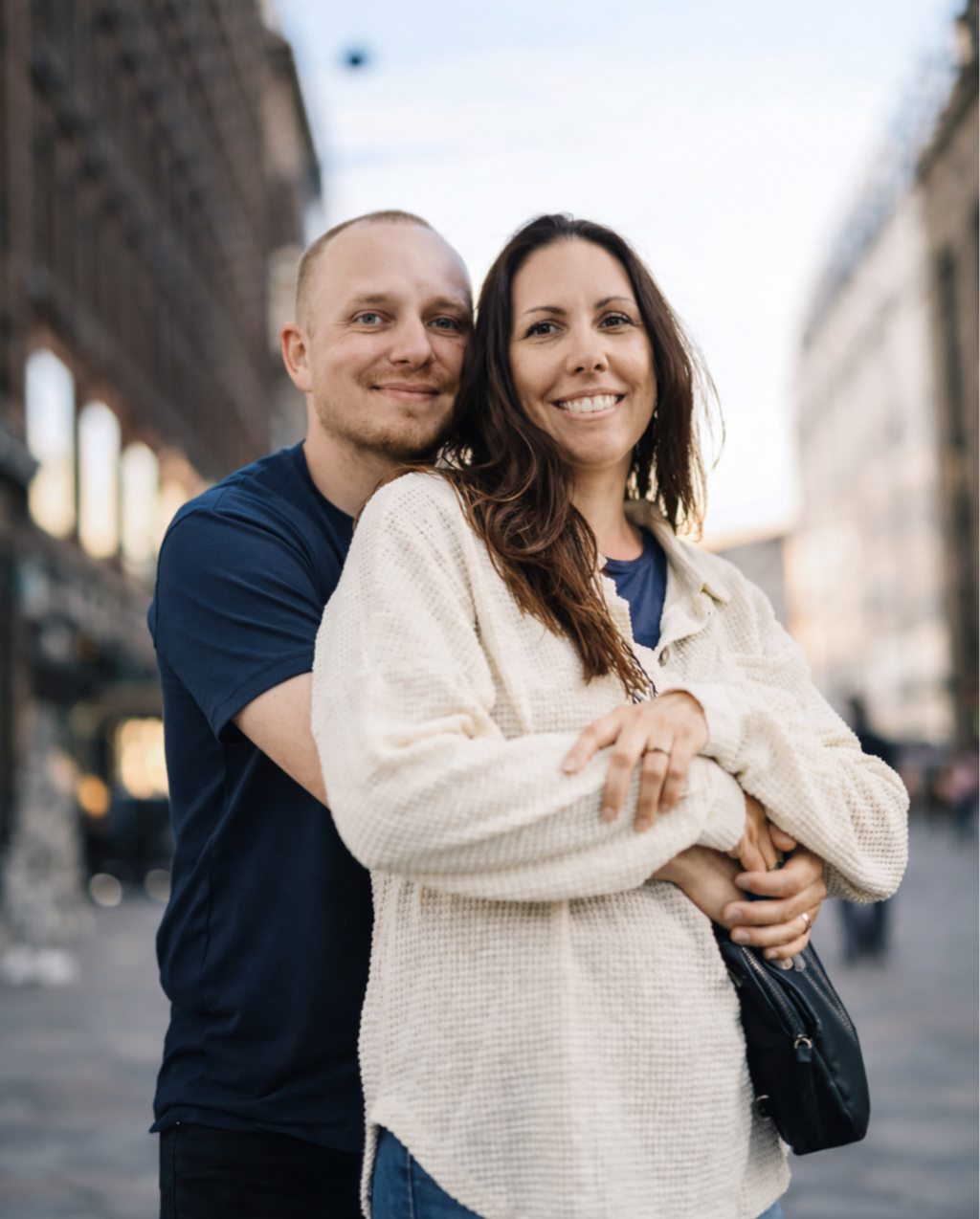 A smiling couple embracing on a city street during the daytime, with buildings and a clear sky in the background.