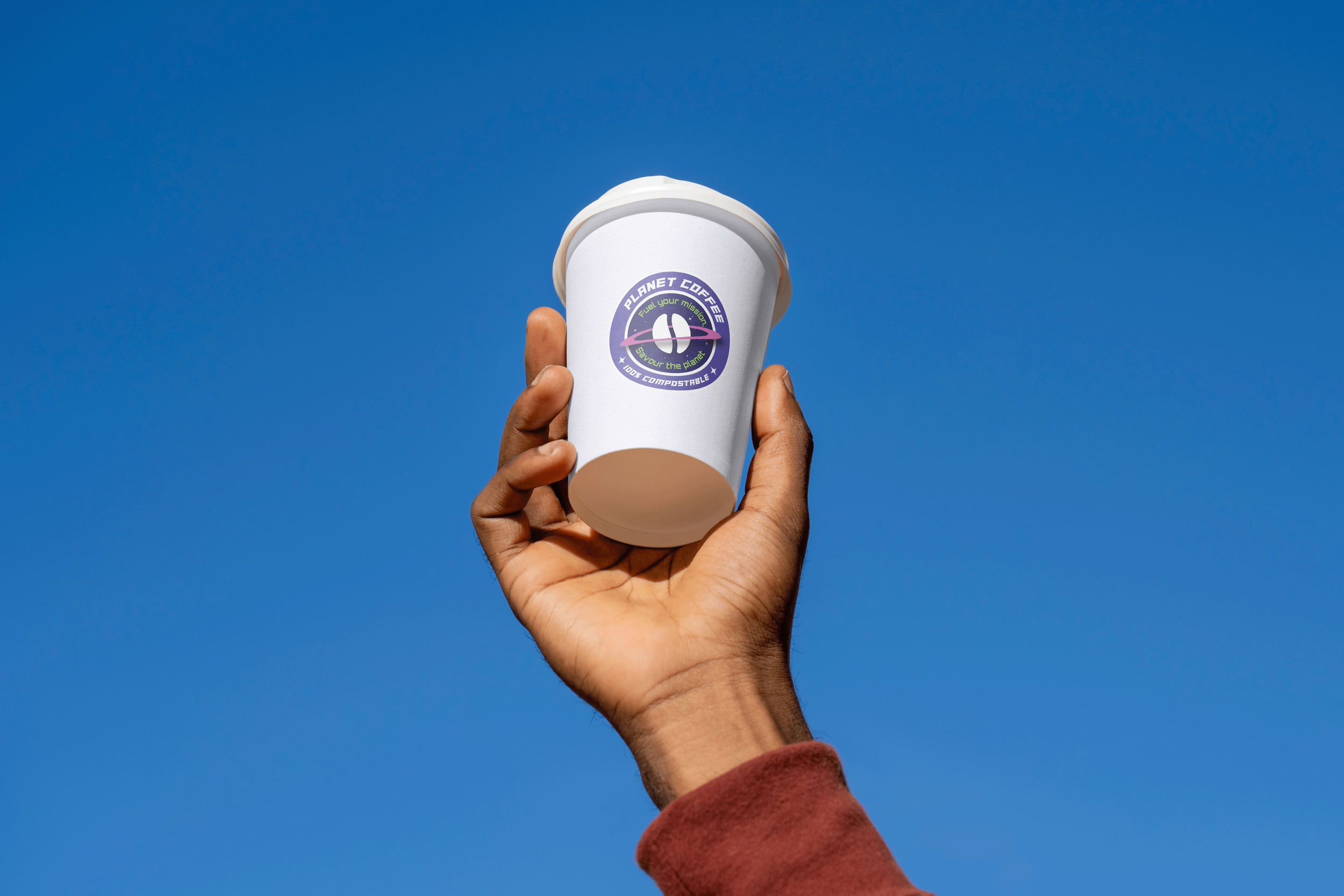 Hand holding a white paper cup with a colorful sticker that reads "Planet Coffee" against a clear blue sky.
