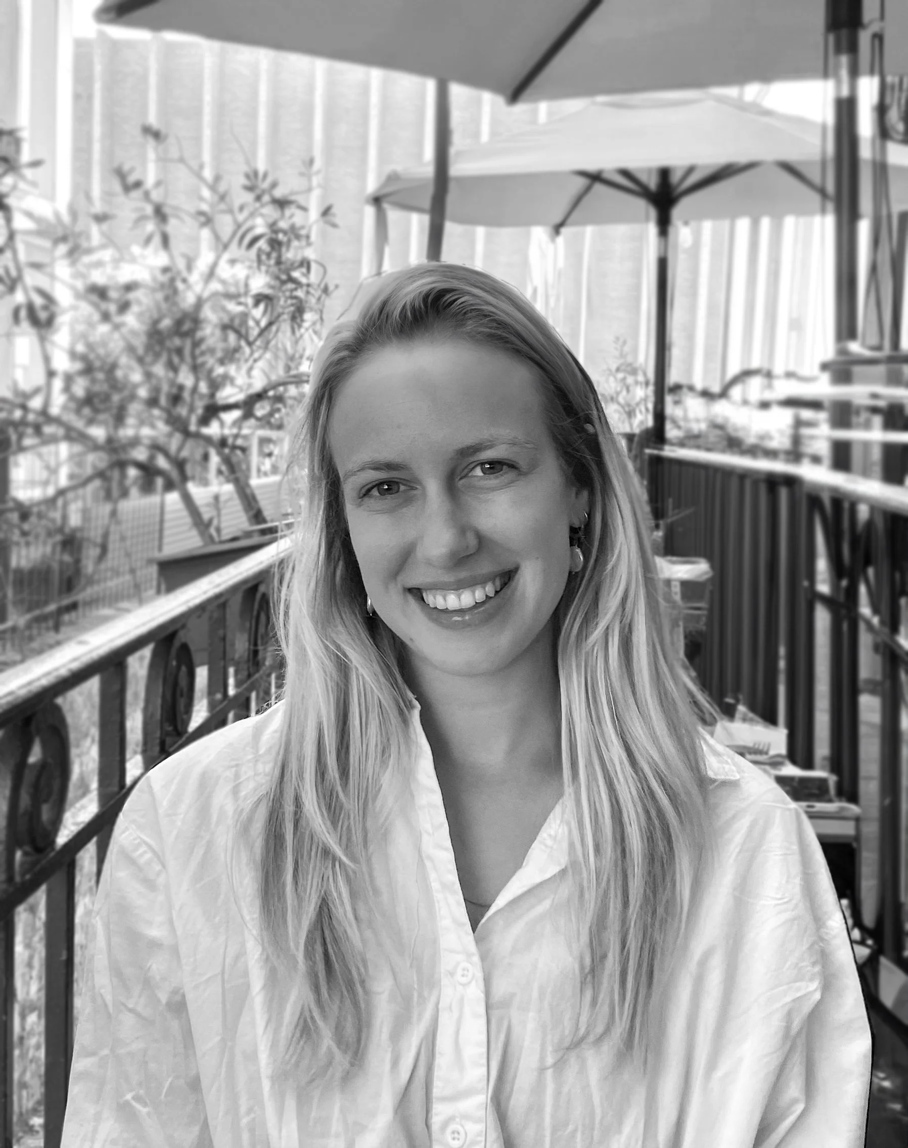 A smiling woman with long blonde hair is wearing a white shirt and is outdoors under large patio umbrellas, with a balcony railing and some trees in the background.