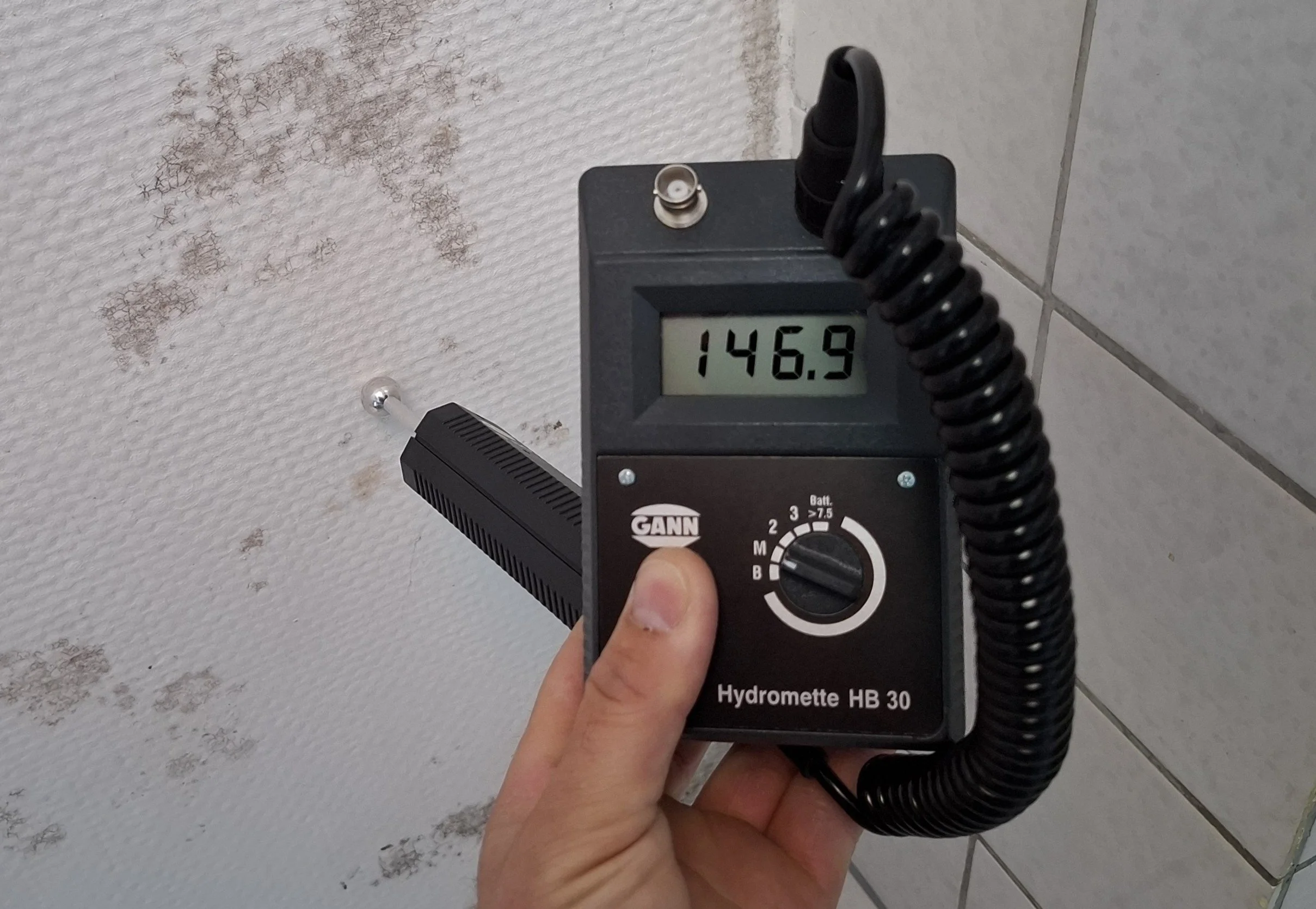 Hand hält ein Hydromete-Gerät vor einem verschmutzten weißen Wand.