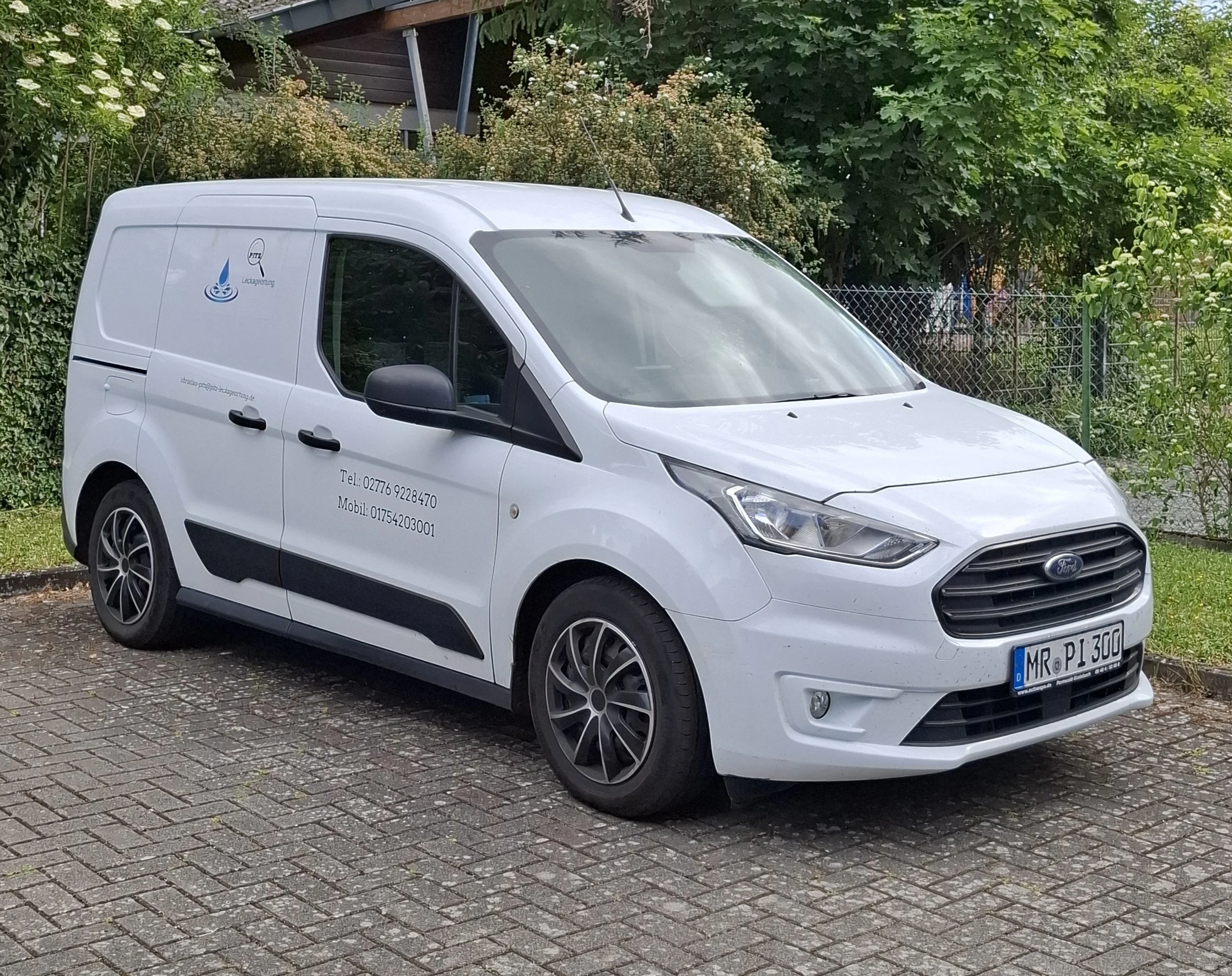 Ein weißer Lieferwagen mit Firmenlogo und Kontaktdaten parkt auf einem gepflasterten Parkplatz, umgeben von Bäumen und Grünflächen.