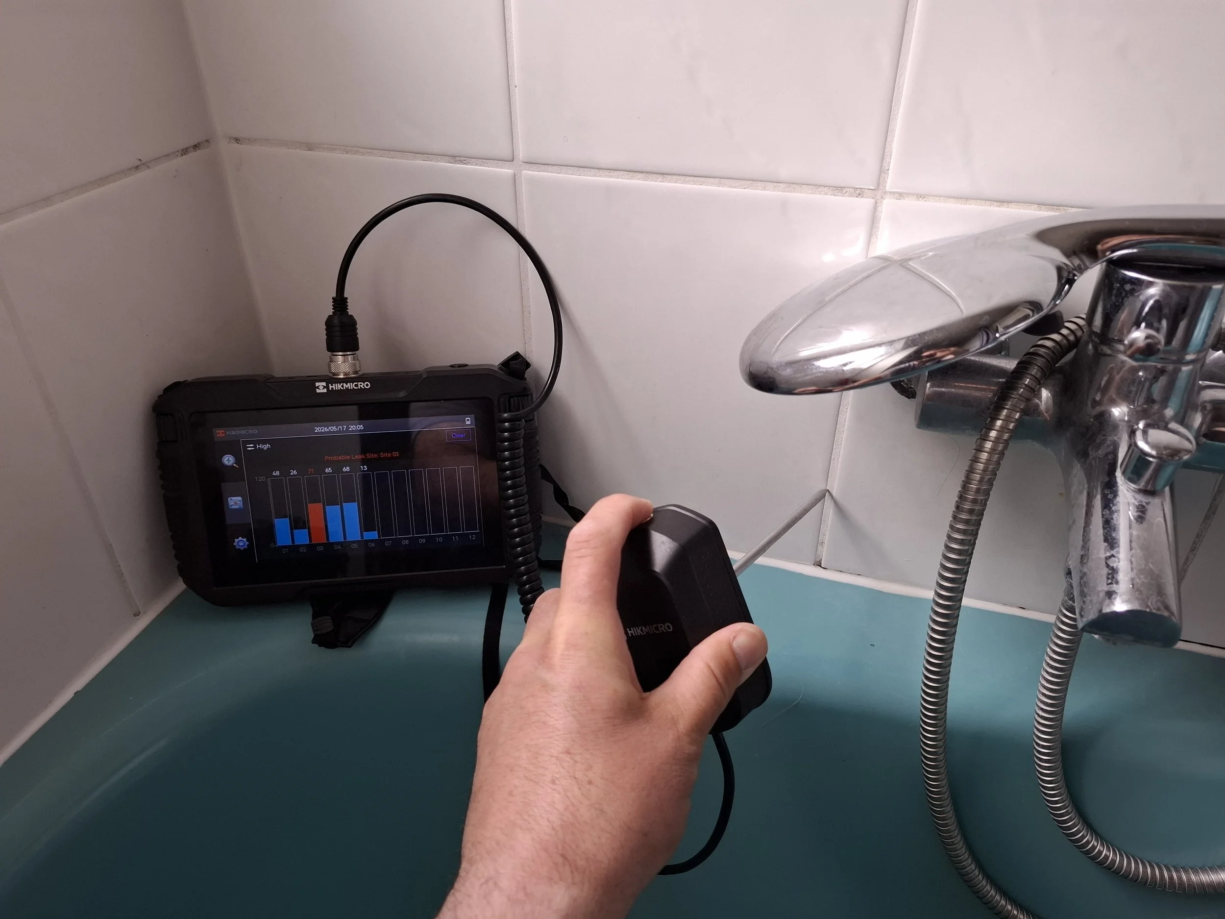 Ein Messgerät mit digitalem Display, das auf einer weißen Fliese liegt, verbunden mit mehreren Kabeln, einer Person hält ein Kabel in der Hand.