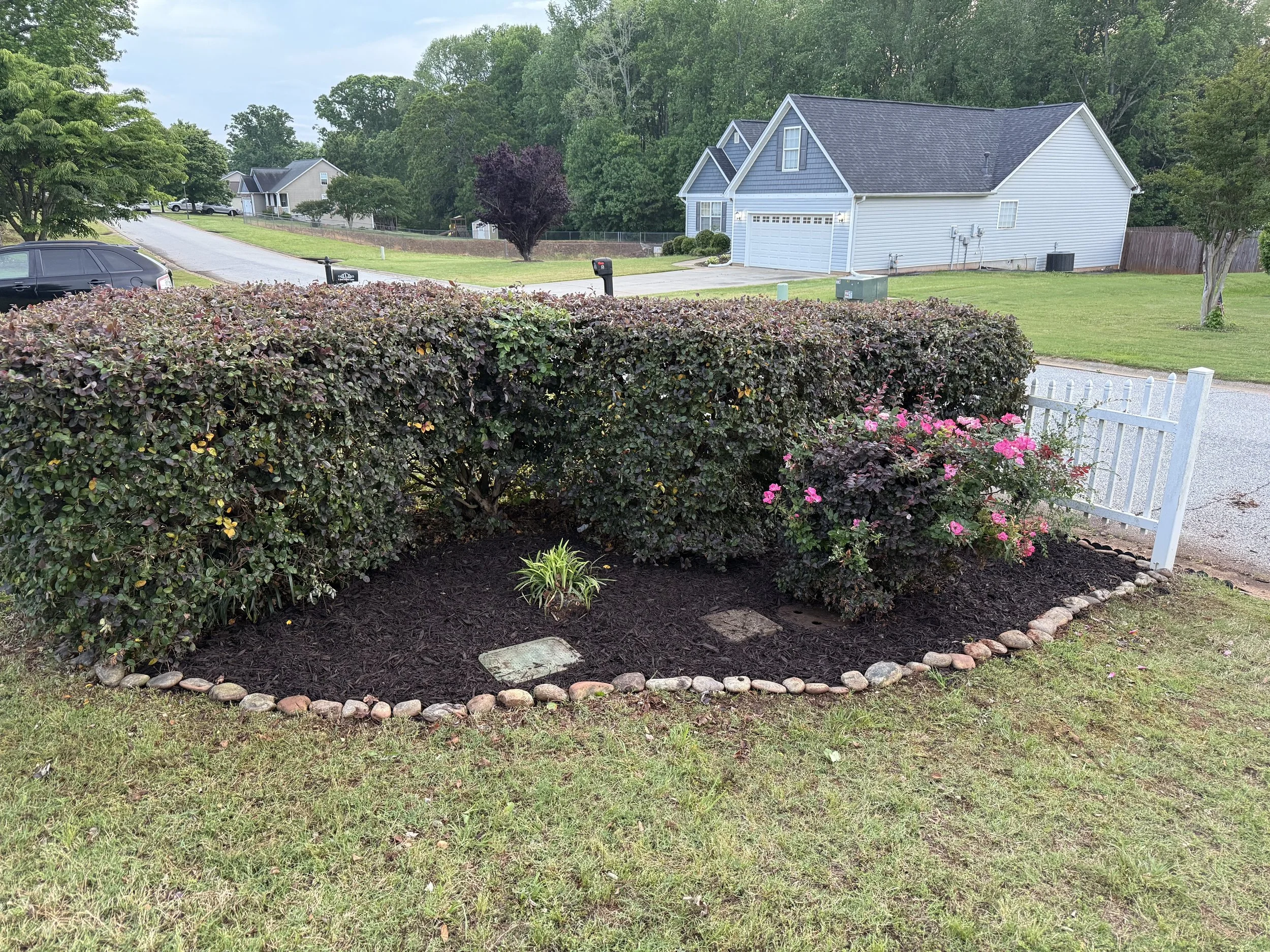Neighborhood scene with a white house, green lawn, trees, a black car parked on the driveway, and a garden bed with pink flowers surrounded by stones at the front.