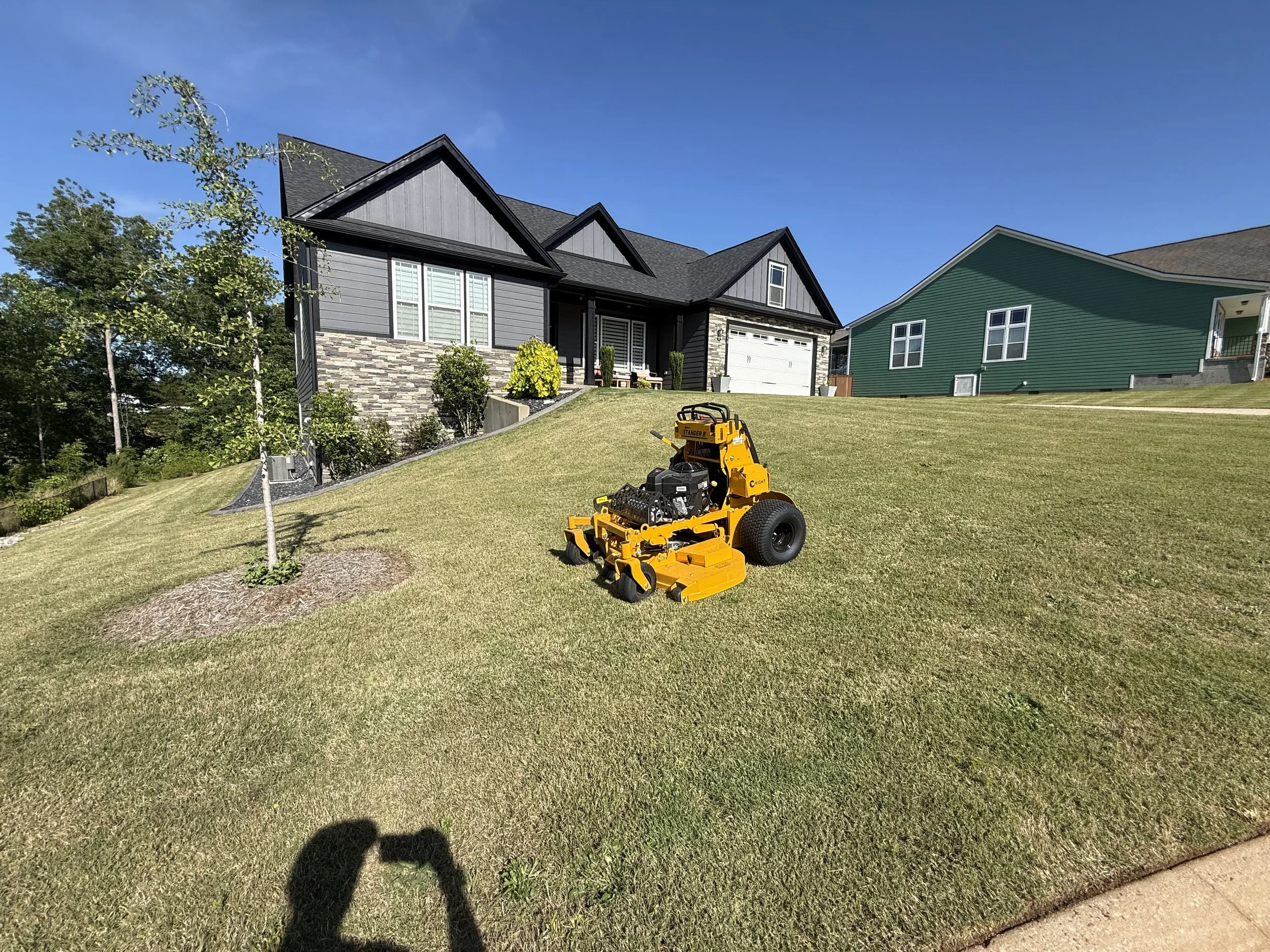 Photo of a yellow commercial lawn mower on well-maintained grass in front of suburban houses with trees and a clear blue sky.