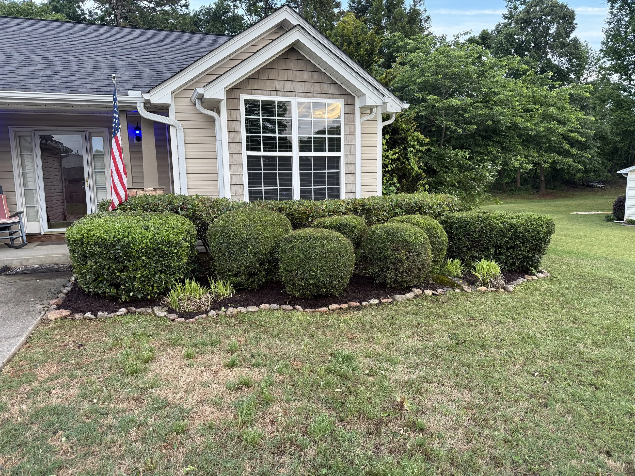 Front of a house with a large window, bushes, and a flag on the porch, surrounded by green trees and a lawn.