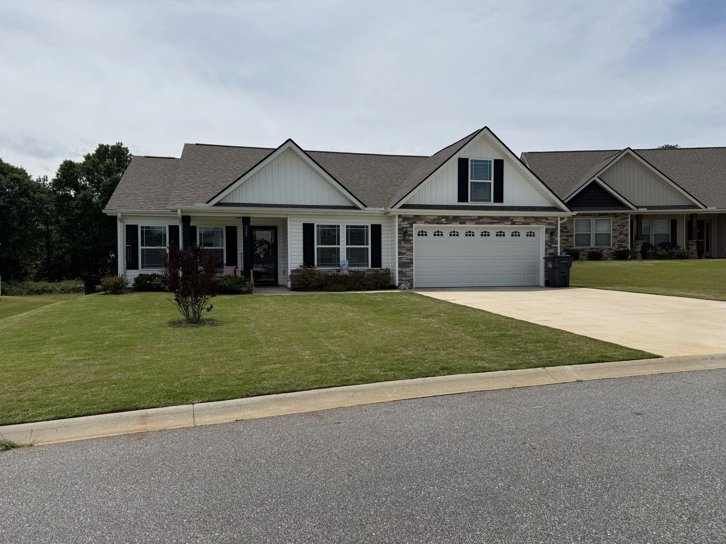 A suburban house with a gray roof, white porch, and a two-car garage with a white door. Well-kept lawn with a small tree and shrubs, street curb, and a concrete driveway.
