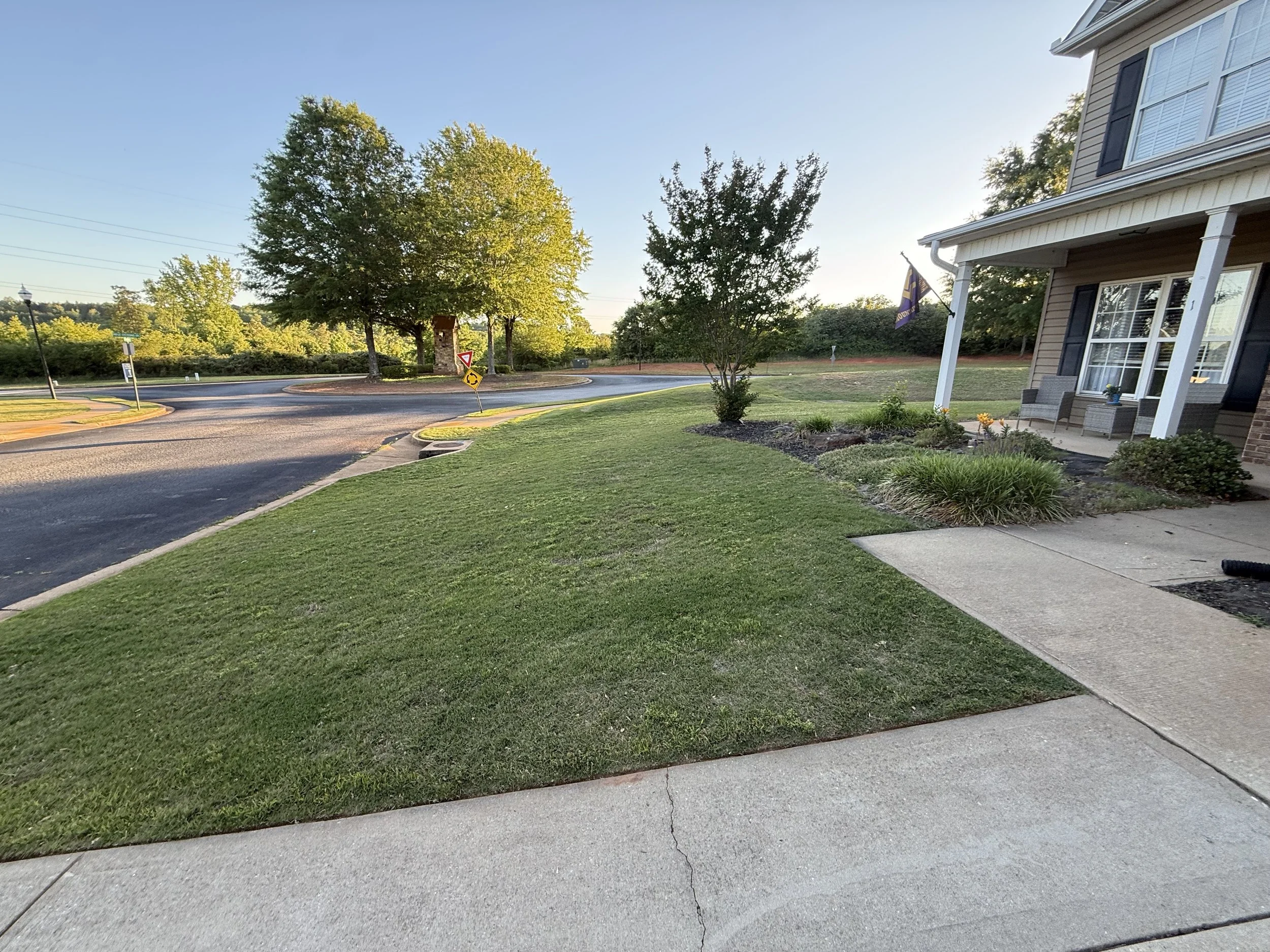 View of a suburban neighborhood with a well-maintained lawn and sidewalk in the foreground, a two-story house on the right, trees and a roundabout with traffic signs in the background, and clear blue sky.