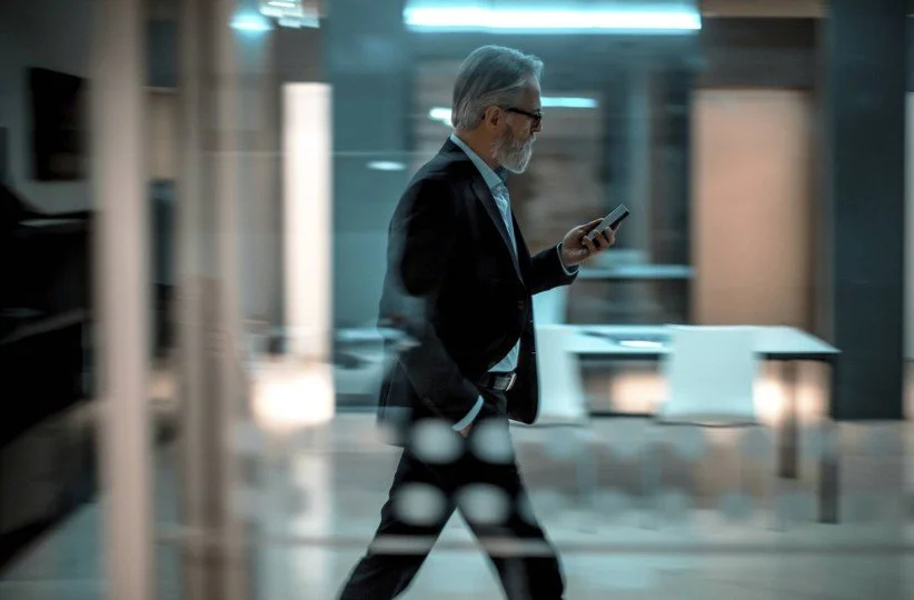 A person with glasses, dressed in a suit, walking and looking at a smartphone in a modern office.