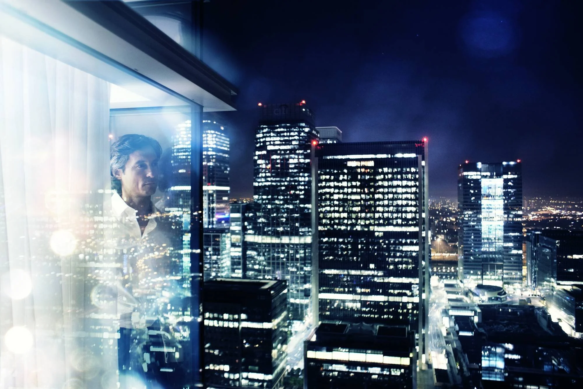 Night-time city skyline viewed through a large glass window, reflecting a person wearing a white shirt.