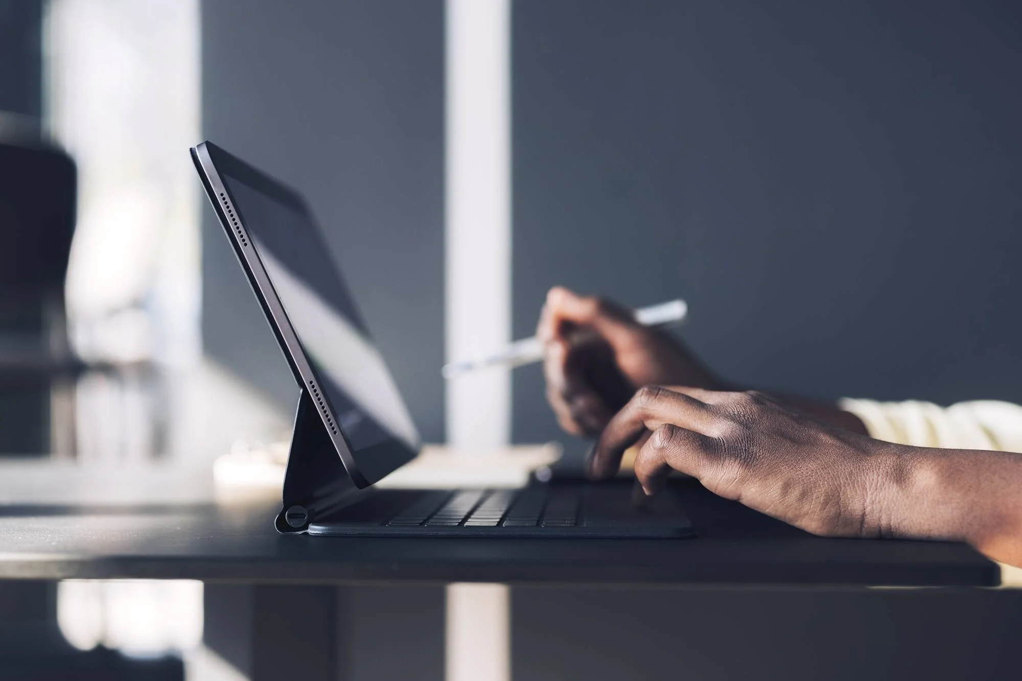 Close-up of a person using a laptop with their hand on the keyboard and holding a stylus pen, with a tablet propped up in front of them on a desk in a modern office environment.