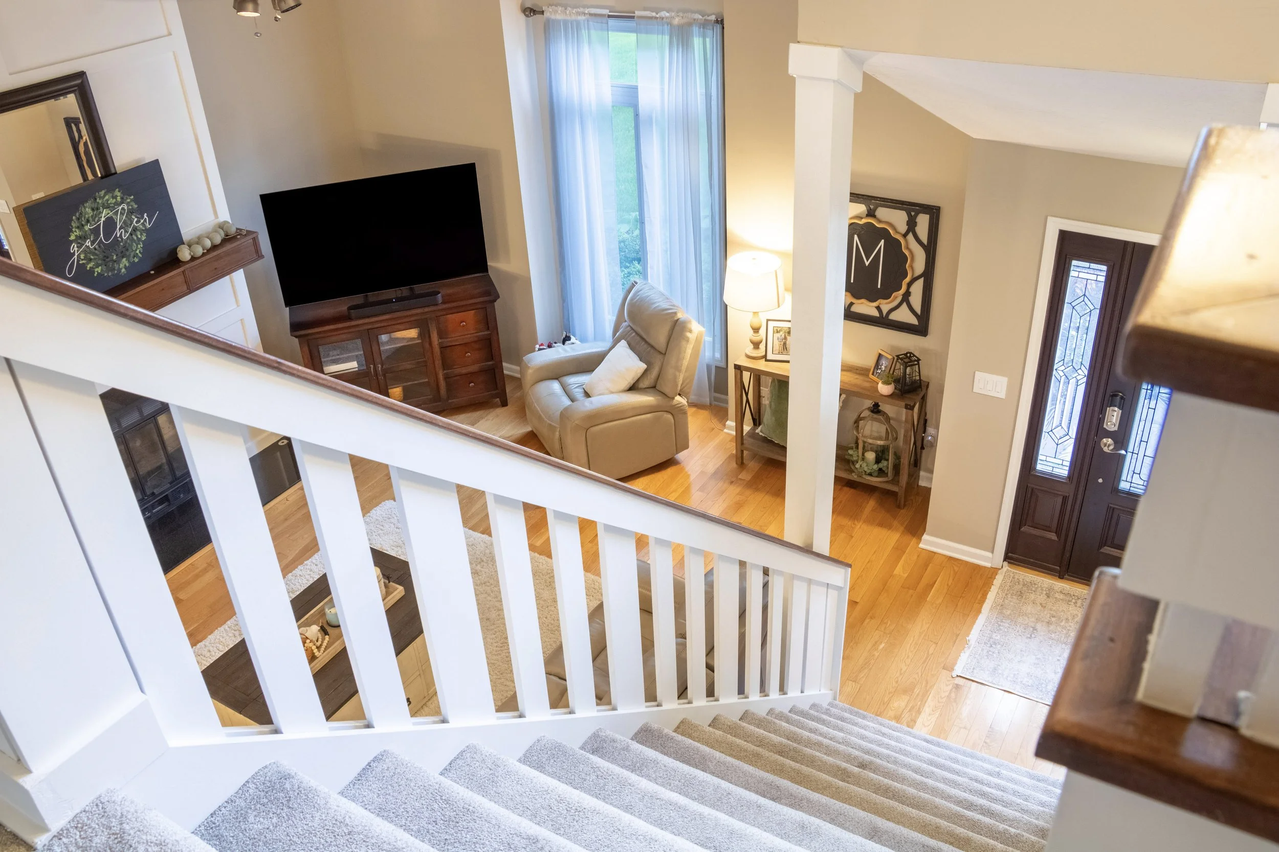 Interior view of a living room seen from the top of a staircase. The room has a beige armchair, a wooden TV stand with a large flat-screen TV, a side table with a lamp, decorative framed art on the wall, and a large window with sheer curtains letting