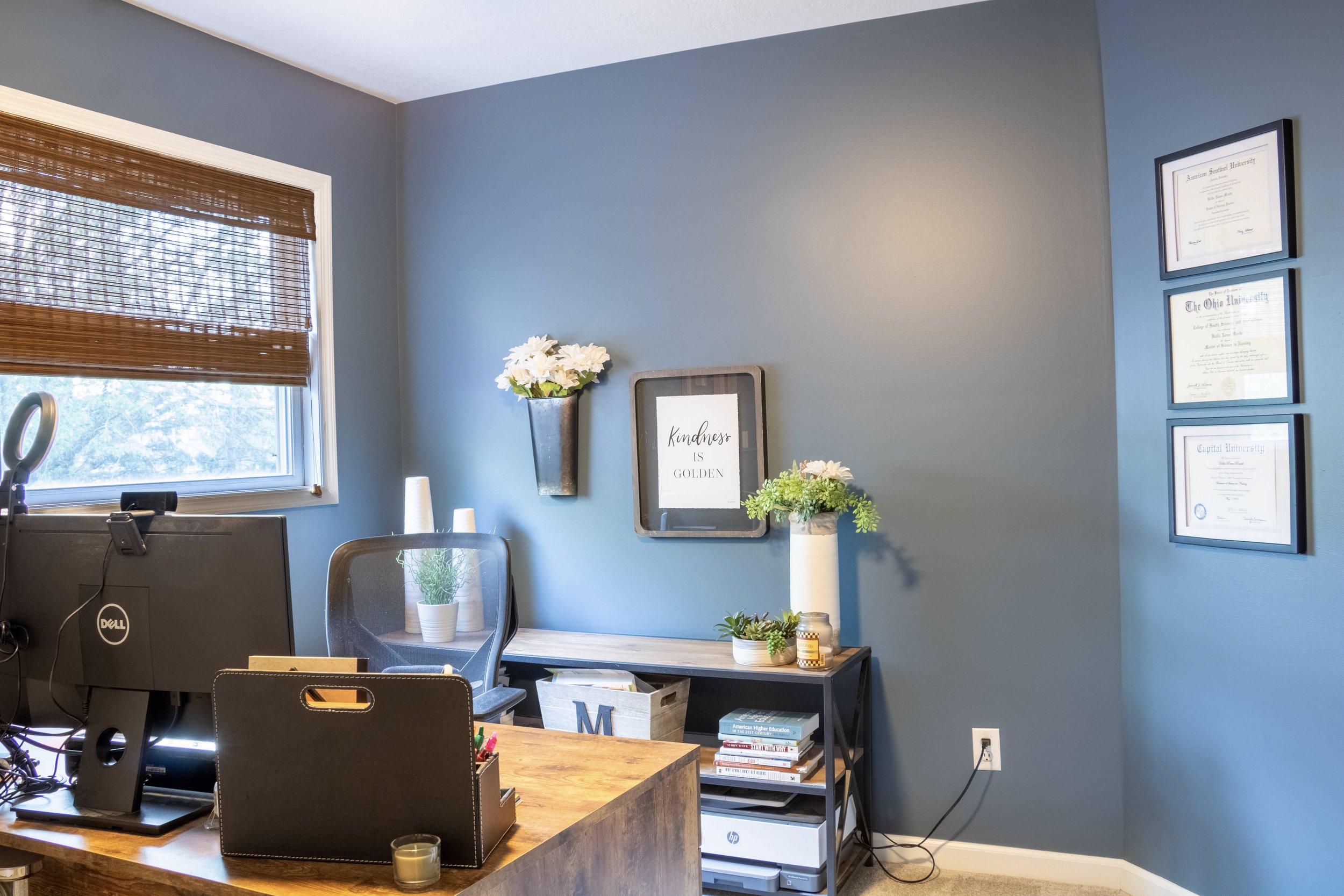 An office corner with a wooden desk, a gray chair, a window with bamboo blinds, and framed university diplomas on a blue wall. Decor includes a potted plant, a framed quote, and a vase with white flowers.