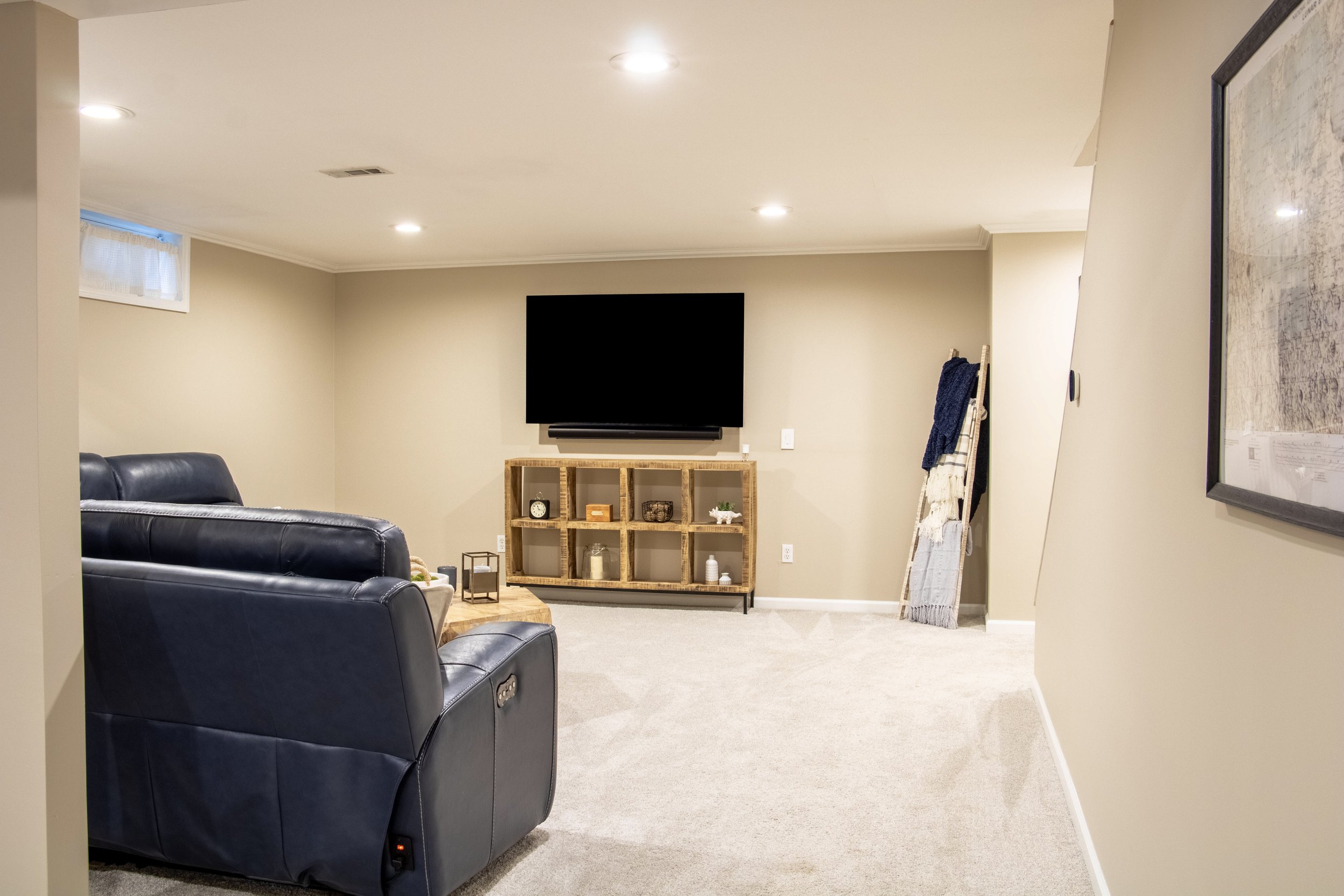 A cozy basement living room with beige walls and carpeted floor. It features two black leather recliners, a wooden shelving unit with decorative items, a wall-mounted flat-screen TV, and a small window with a white curtain. There is a ladder with tow