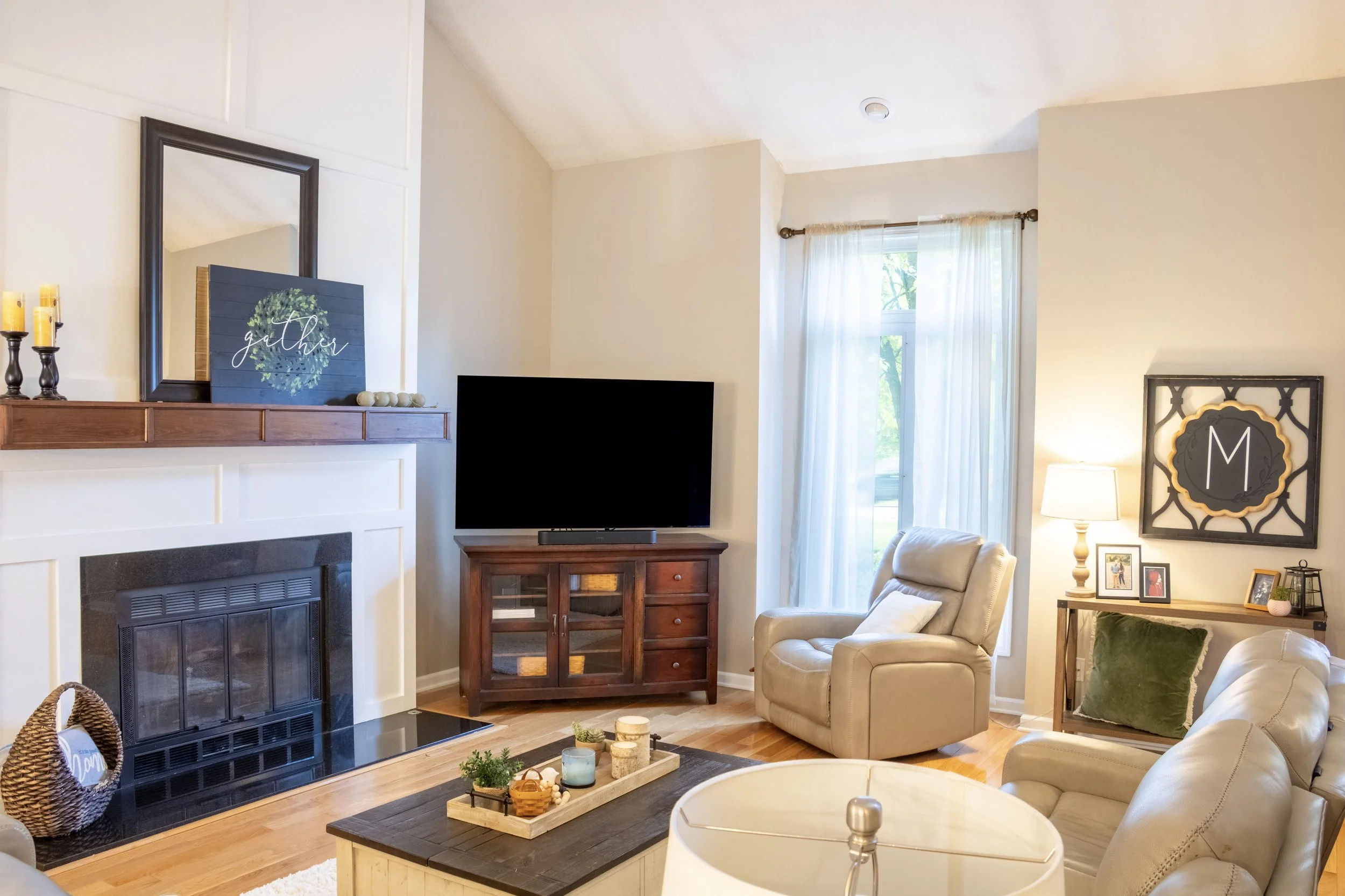 Living room with beige walls, white curtains on large windows, a white armchair, a wooden cabinet with a TV, and decorative items including a black-framed mirror, a sign that says 'gather' with a green wreath, and framed photos on a side table.