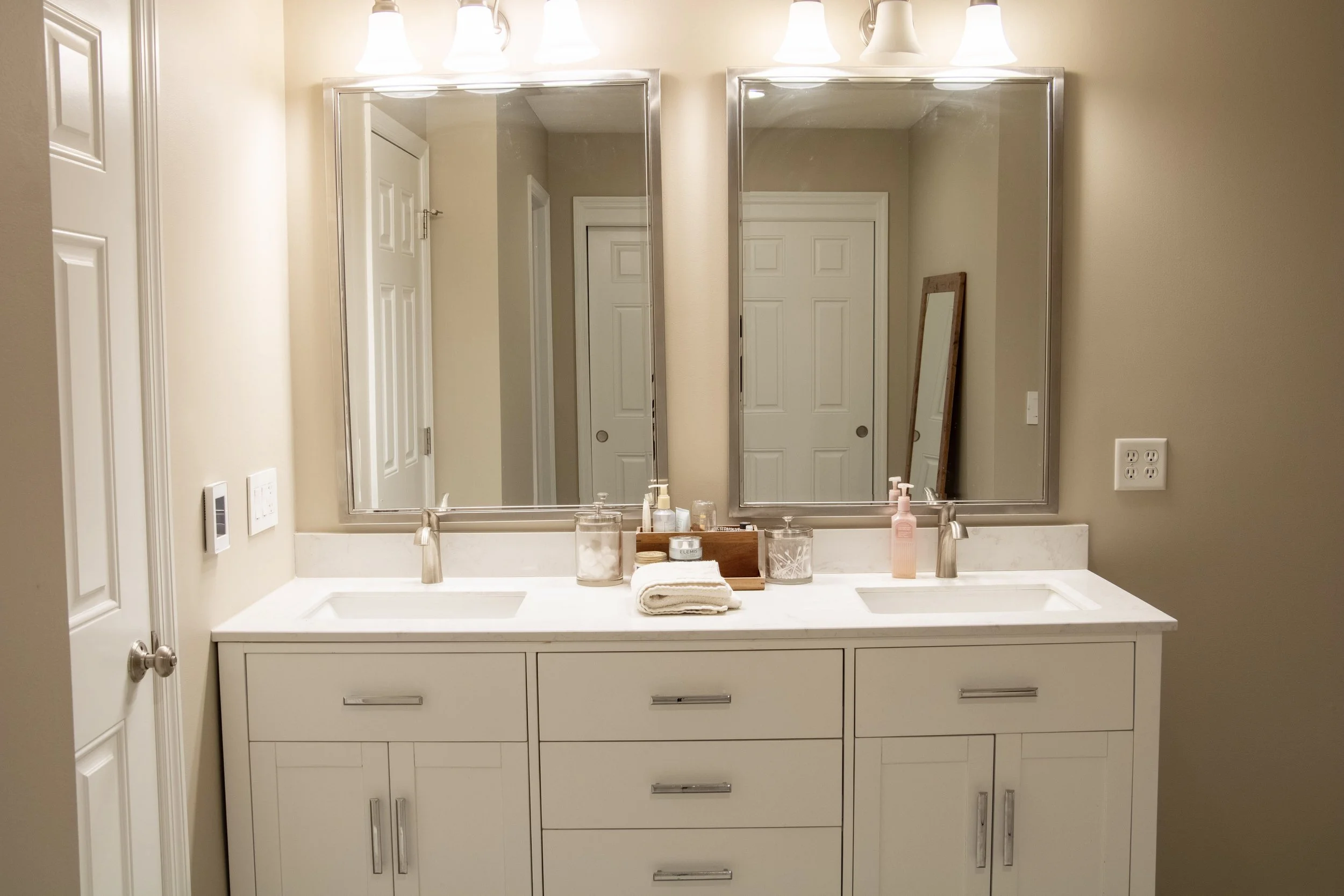 Bathroom vanity with two sinks, large mirror, and wall-mounted light fixtures. Items on the countertop include towels, soap dispensers, and toiletries. Reflection shows a door and a picture frame on the wall.