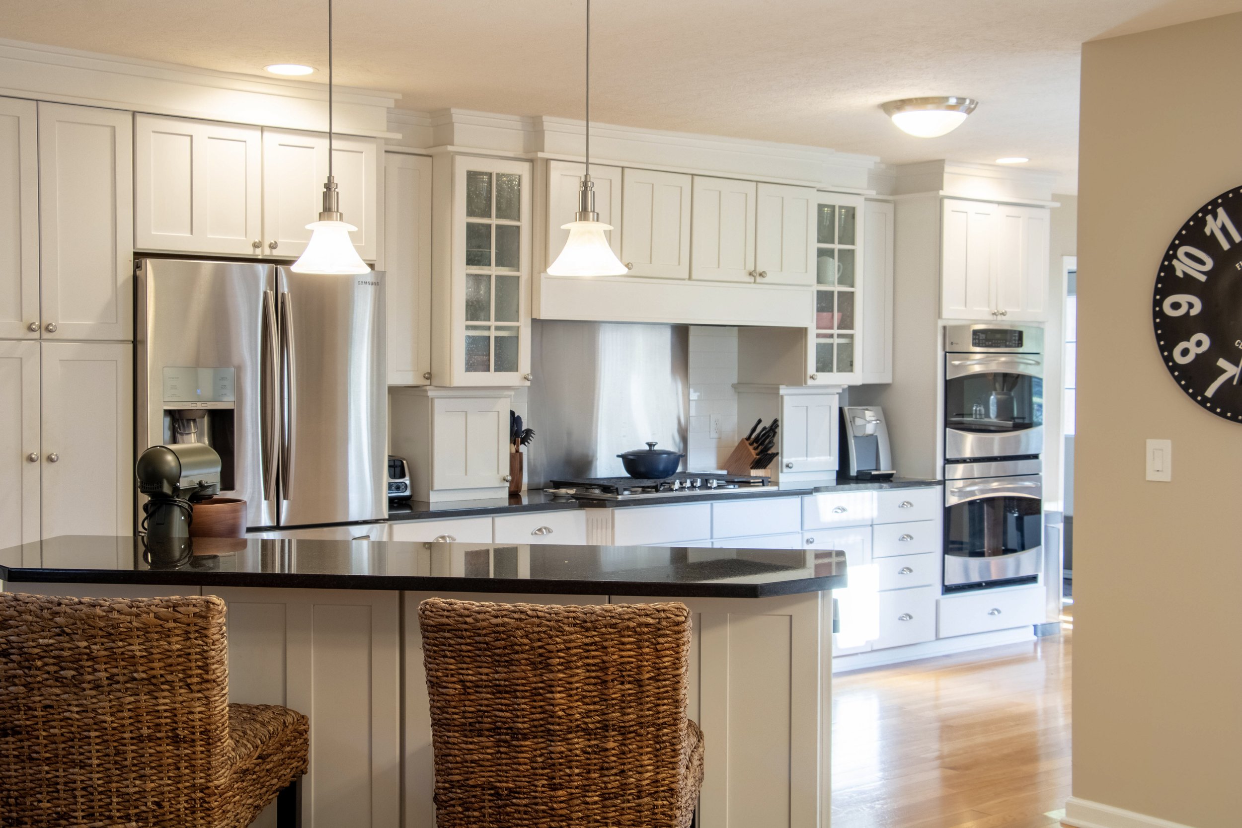 A kitchen with white cabinets, stainless steel refrigerator, black countertop, and woven rattan chairs at a counter. Pendant lights hang from the ceiling, and there is a wall clock on the right side.
