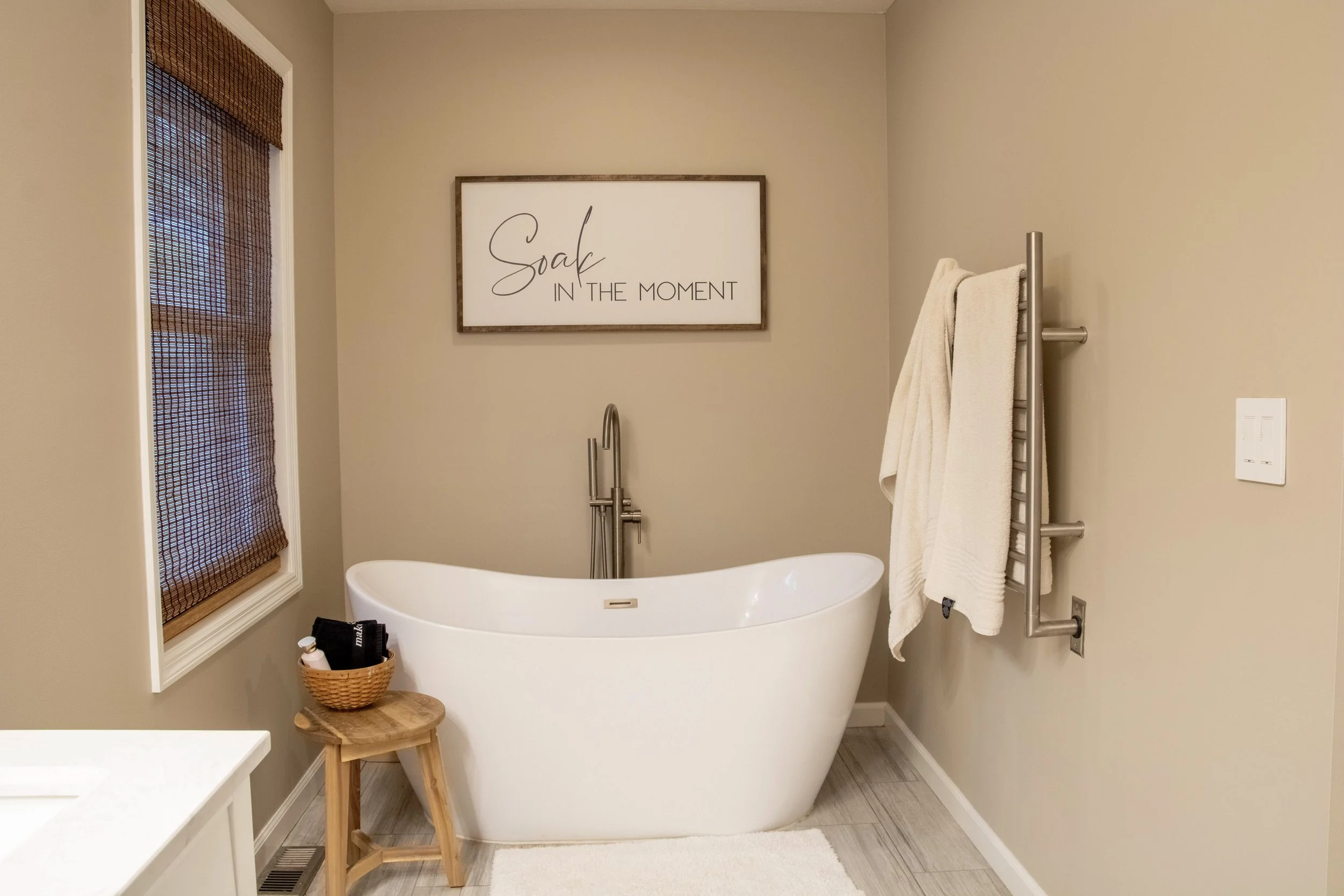 A modern bathroom with a white freestanding bathtub, a wooden stool with items on it, a basket, a towel rack with towels, a window with bamboo blinds, and a framed quote on the wall that says "Soak in the moment."