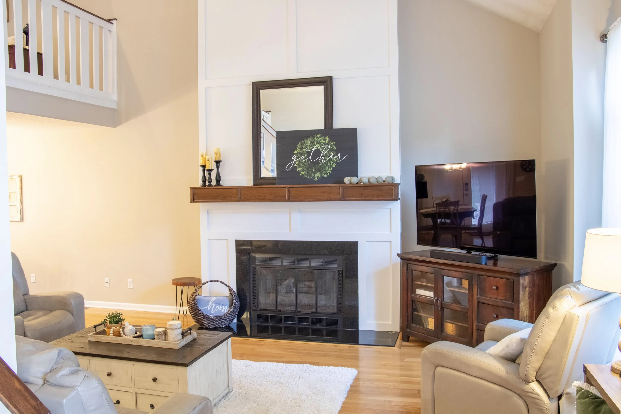 Living room with a fireplace, a flat-screen TV on a wooden stand, a coffee table with decor, and wall decor including a mirror and a black sign that says 'gather' with a green wreath design.