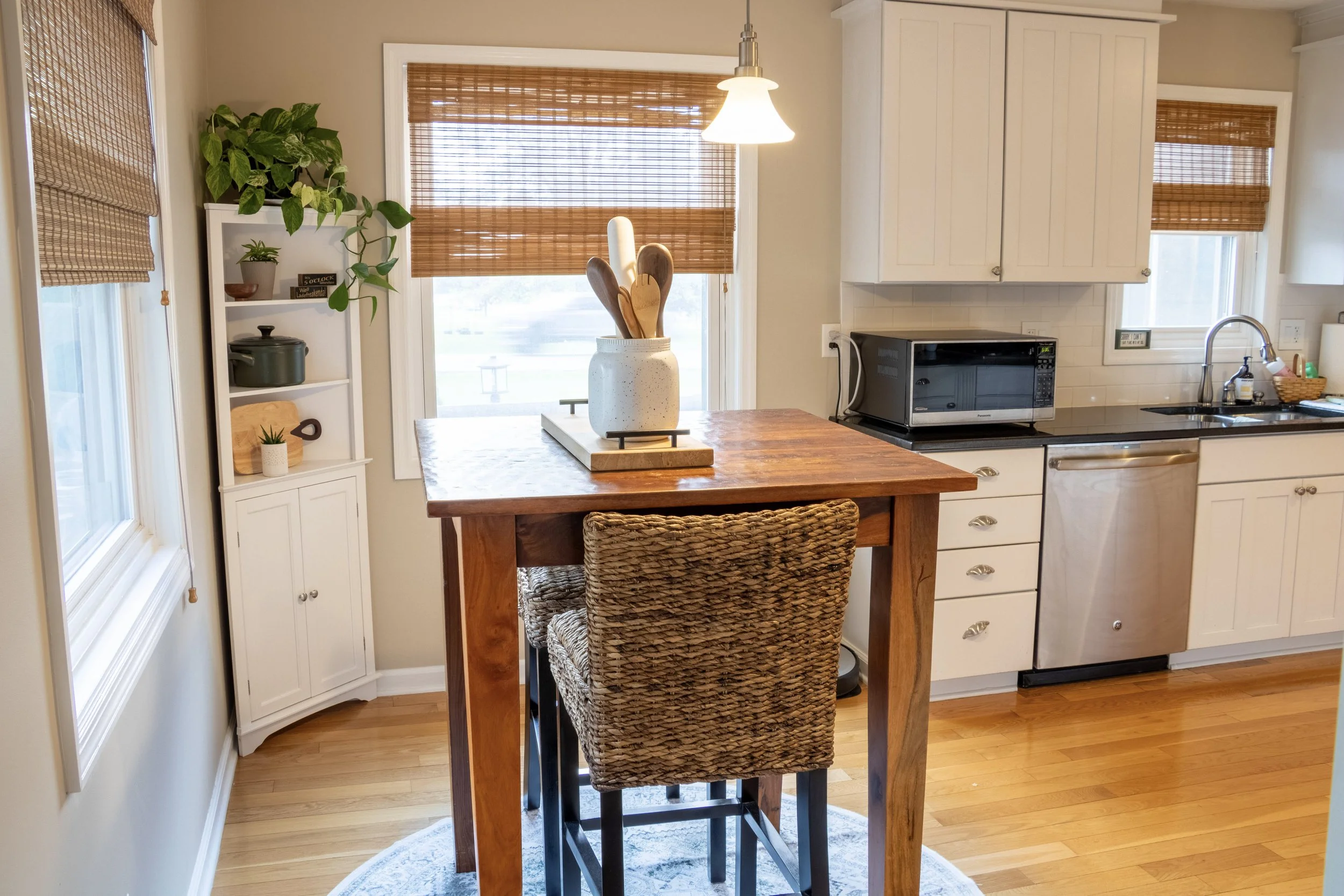 Kitchen with white cabinets, a small wooden table with a wicker chair, a white vase with wooden utensils on the table, window shades, a microwave, a sink with a faucet, and some small plants.