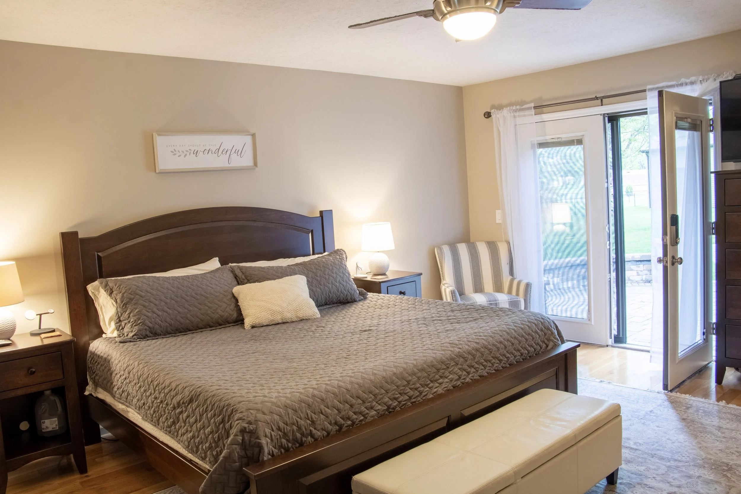 A bedroom with a wooden bed frame, brown quilted bedding, side table with a lamp, striped armchair, and glass door leading outside to a grassy area.