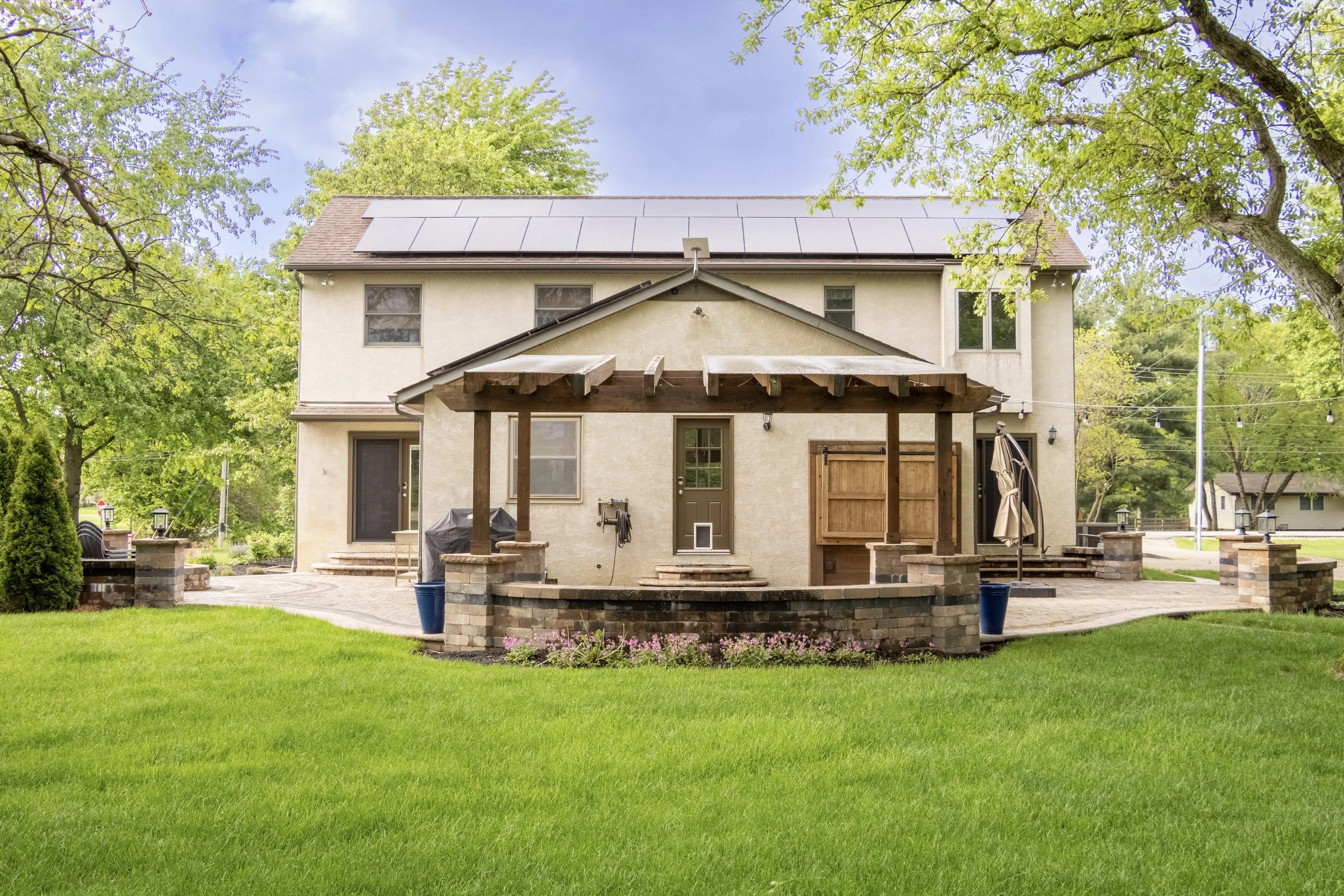 The backyard of a two-story house with beige exterior walls, a deck, and a multi-pane door. The yard has a lush green lawn, a small garden bed with pink flowers, and a paved patio area with a brick fire pit. There is a pergola with wooden beams and t