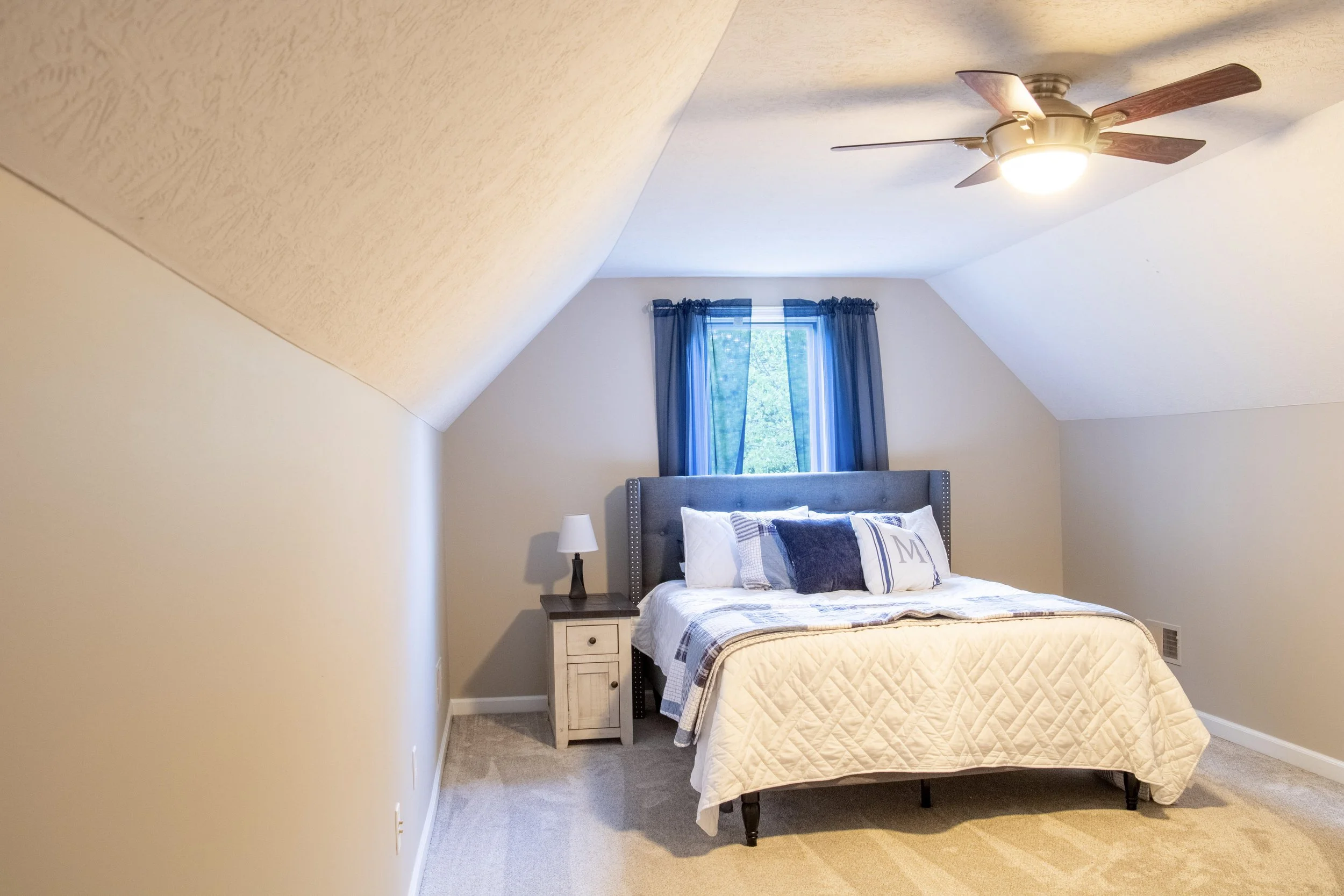 A bedroom with a bed, nightstand, lamp, window with blue curtains, and a ceiling fan with wooden blades.