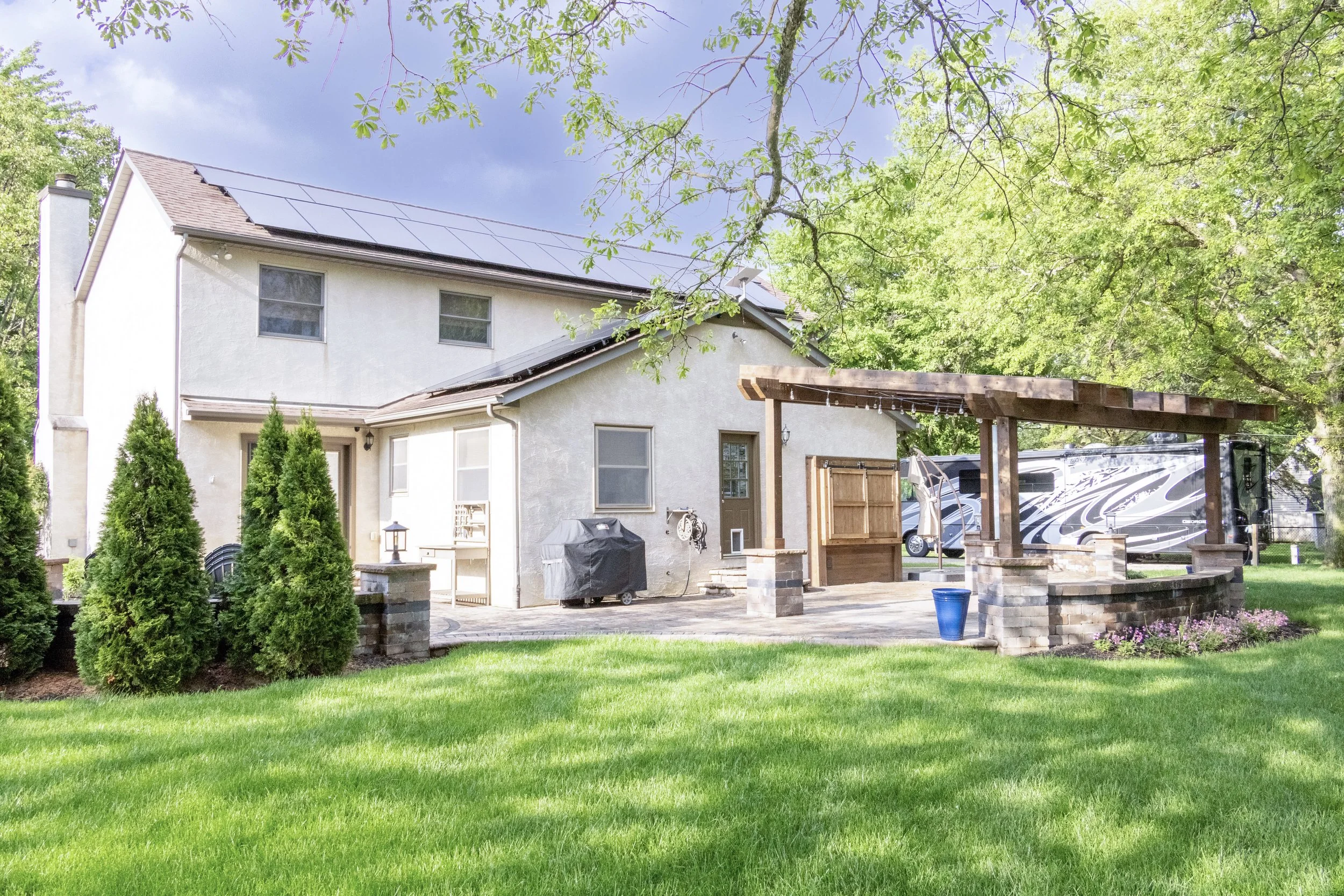 A two-story white house with a brown roof, surrounded by green trees and a well-maintained lawn. There is a covered patio area with a brick and wood structure, a grill, and a RV parked in the background.