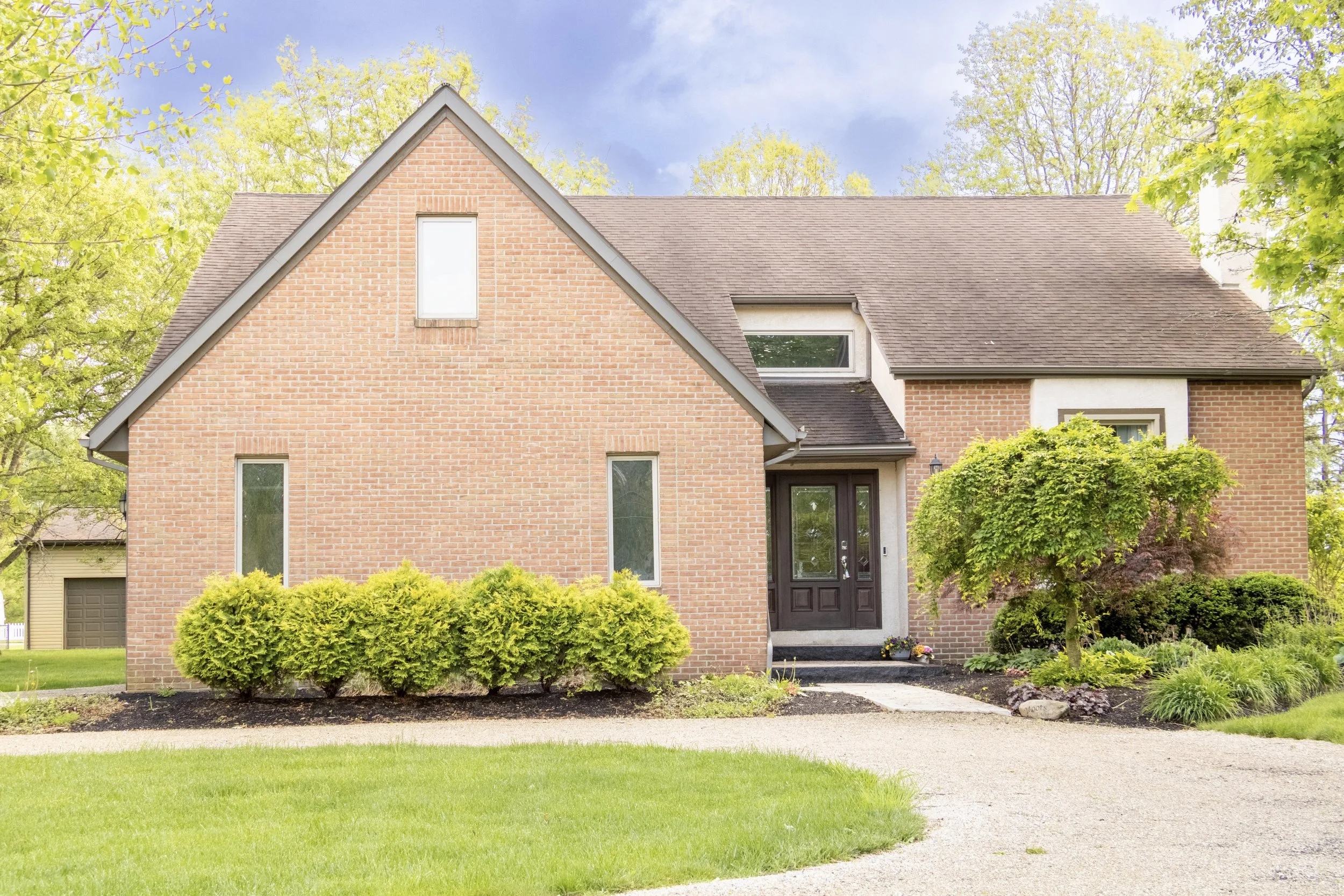 Front view of a brick house with a gabled roof, a black front door, and multiple windows, surrounded by trees and a well-maintained garden with bushes and grass.
