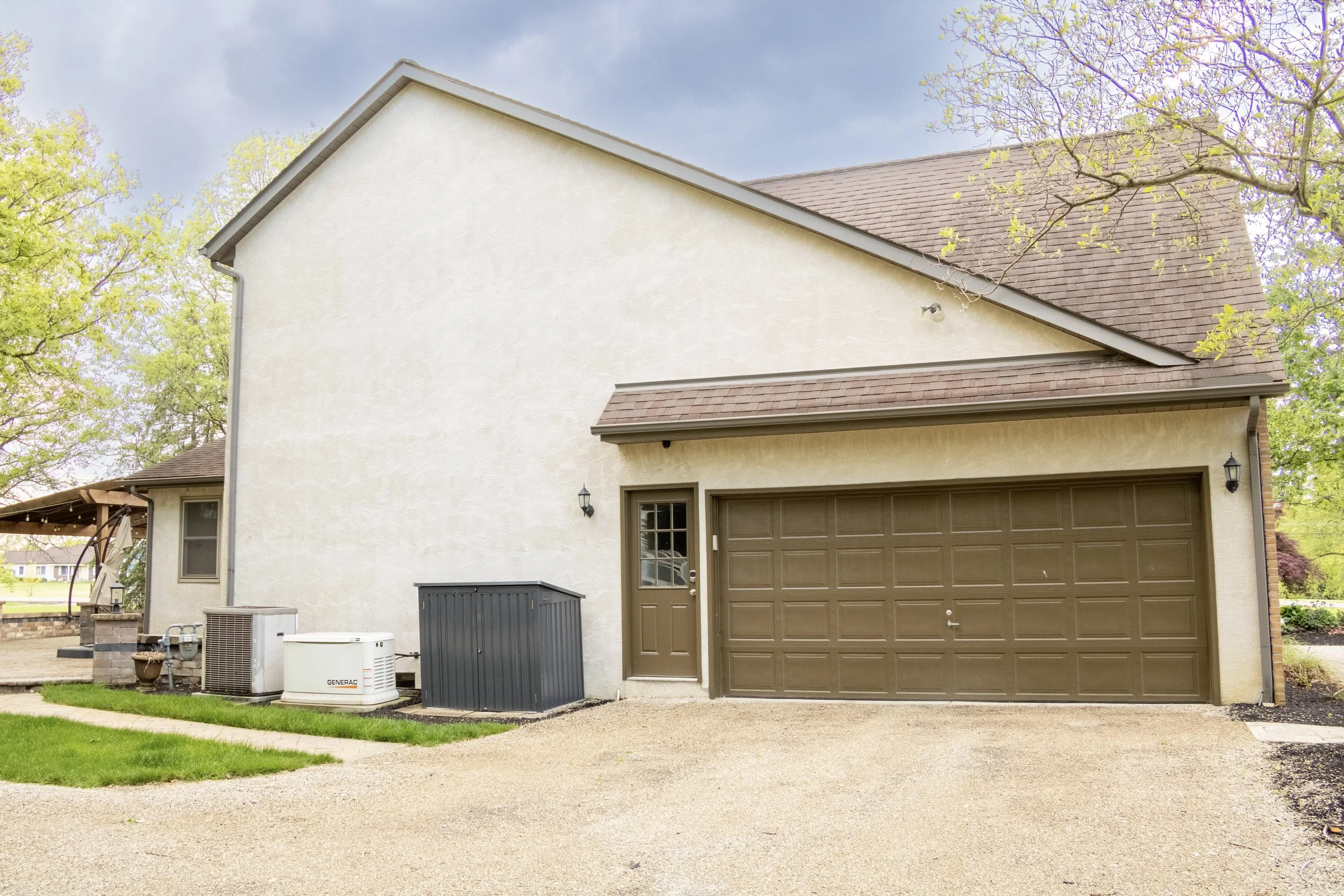 Front view of a house with a beige exterior, brown garage door, and brown roof, surrounded by trees and a paved driveway.