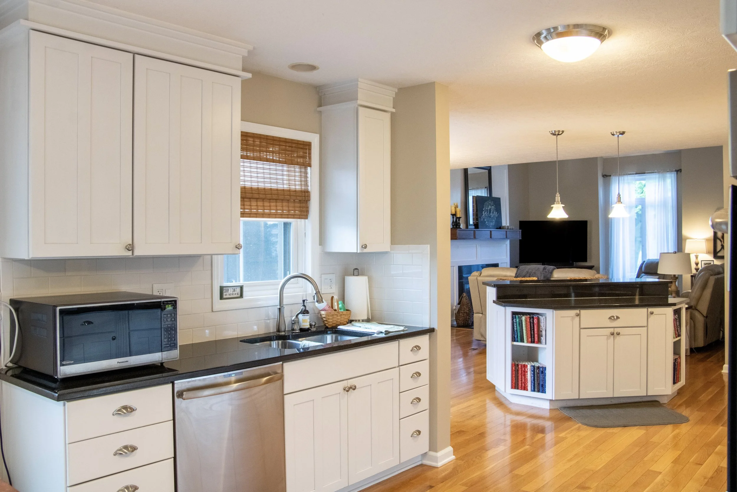 A kitchen with white cabinets, a microwave, and a window with a bamboo shade. In the background, there is an open living room with a television, sofas, and pendant lights.