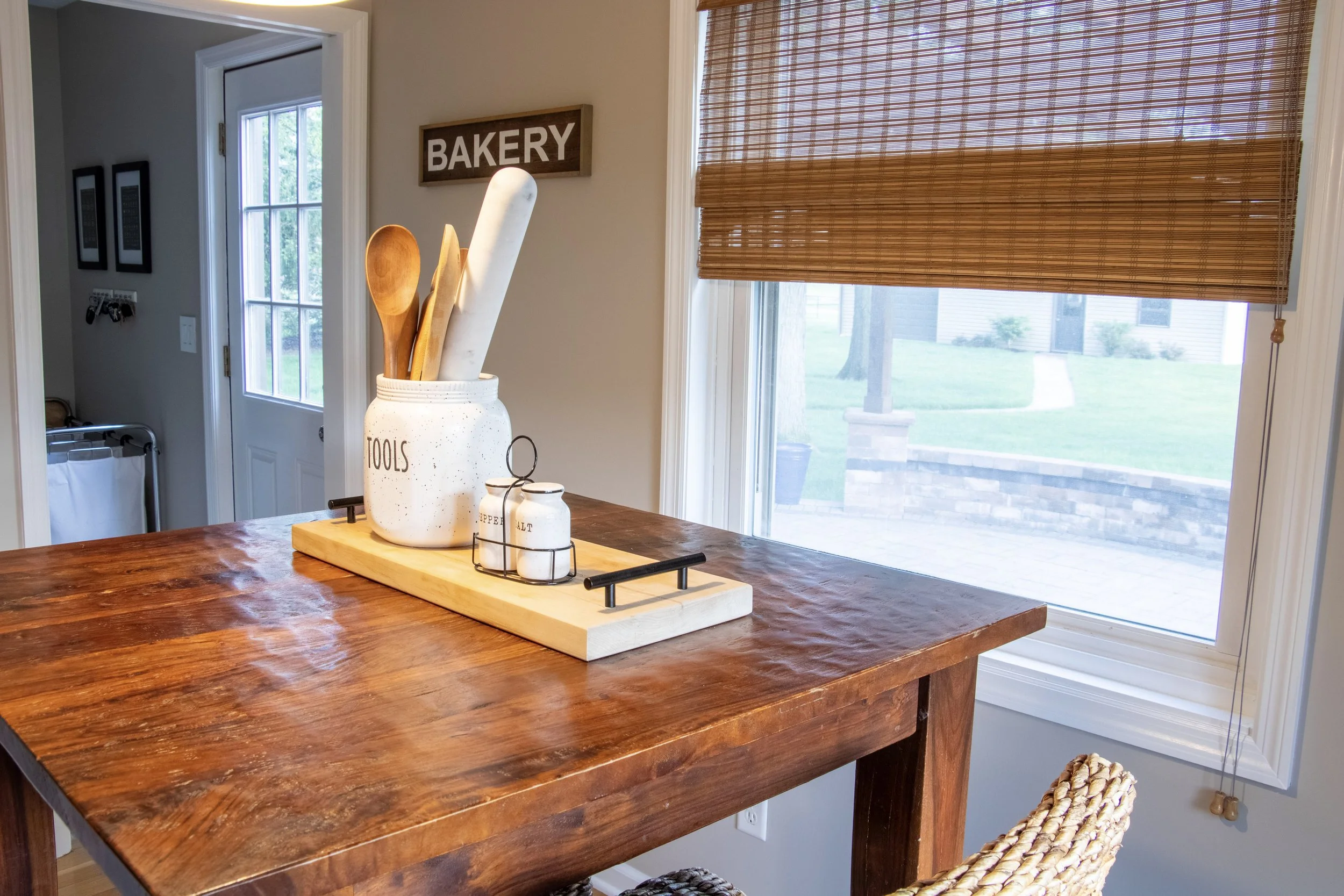 A kitchen or dining area with a wooden table, a white ceramic container holding wooden spoons, and a window with woven bamboo blinds that allows natural light into the room. There is a sign on the wall Reading "BAKERY" and an open door leading outsid