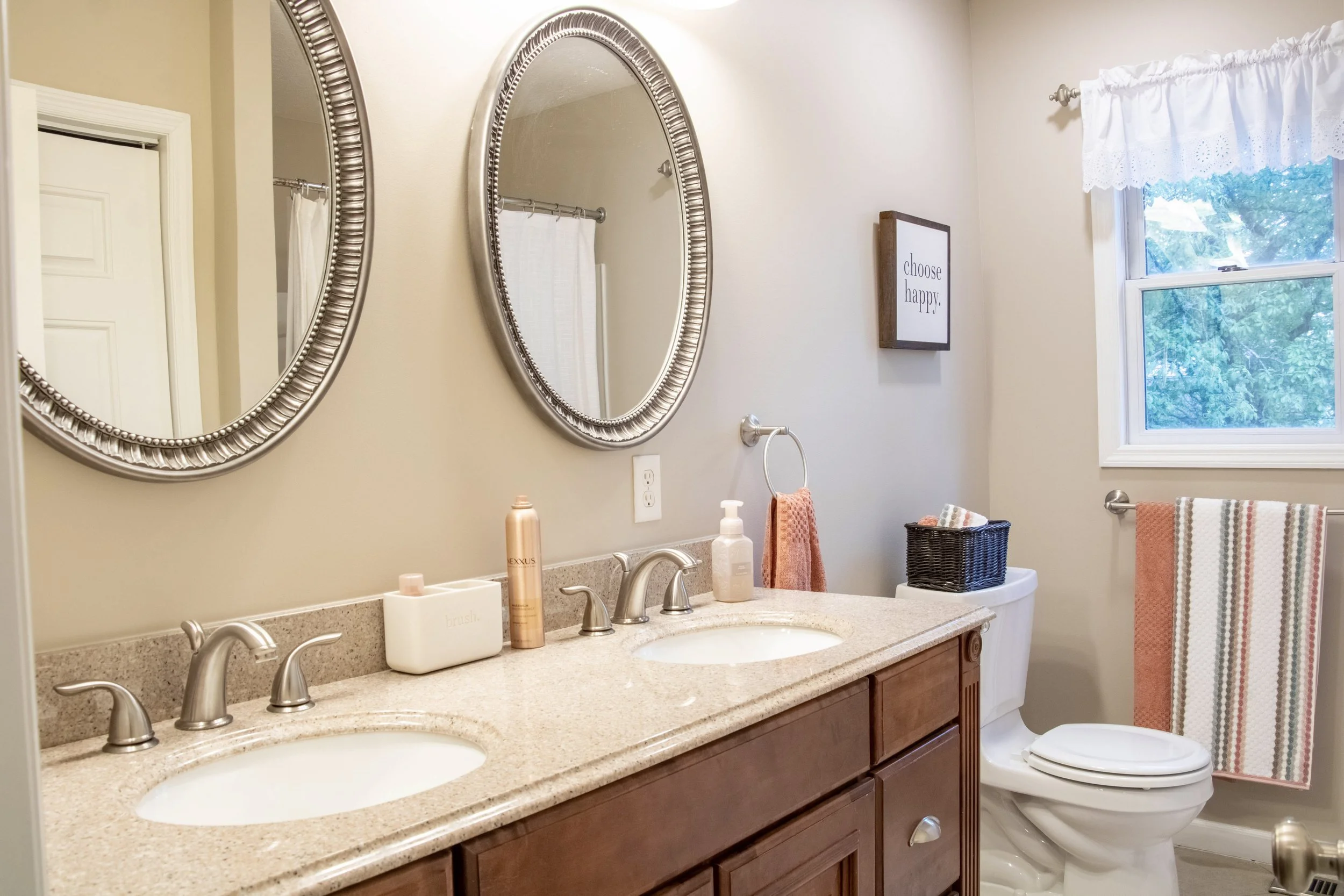 A bathroom with a beige granite countertop, double sinks, and dark wood cabinets. Two large oval mirrors with silver frames hang above the sinks. There are bottles and containers on the counter. A wall-mounted sign reads "choose happy." A window with