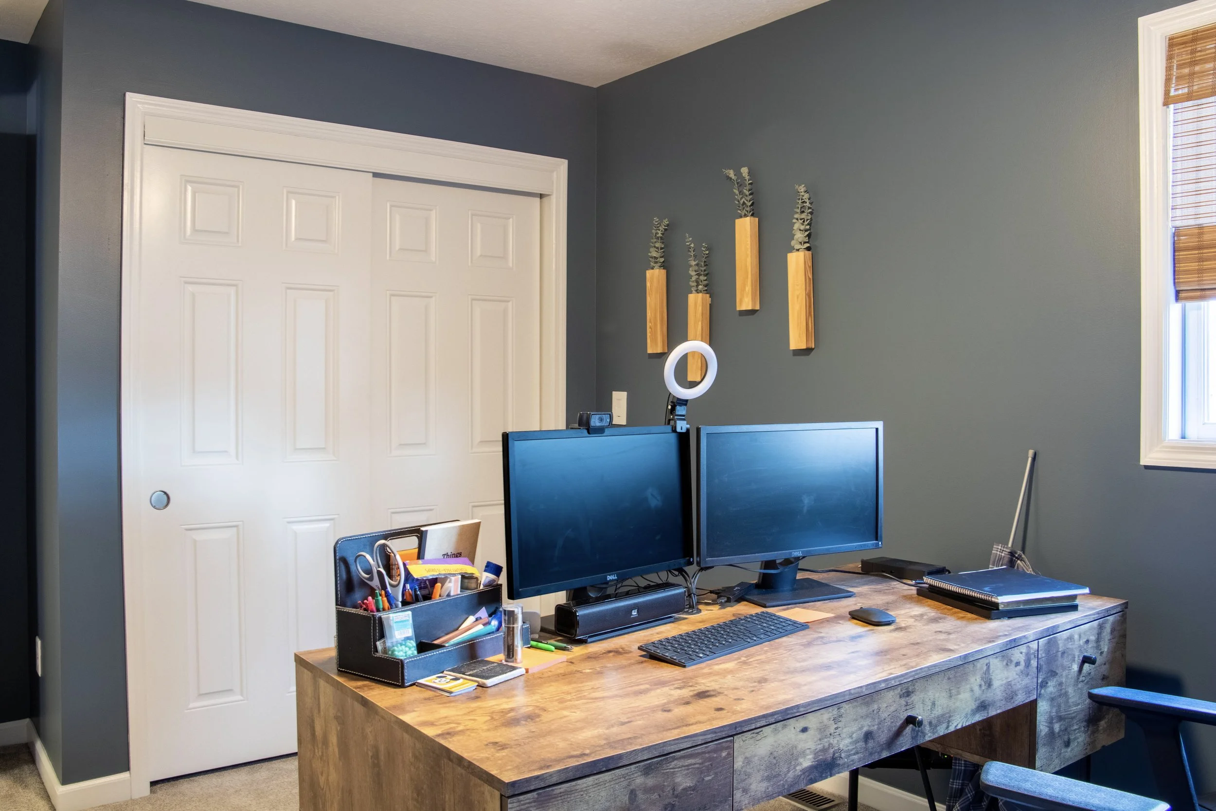 View of a home office with a wooden desk, two computer monitors, a keyboard, a mouse, and office supplies in a container. There are three wooden wall planters with plants on a blue wall, and a white door with a vent in the top panel. A window with ba