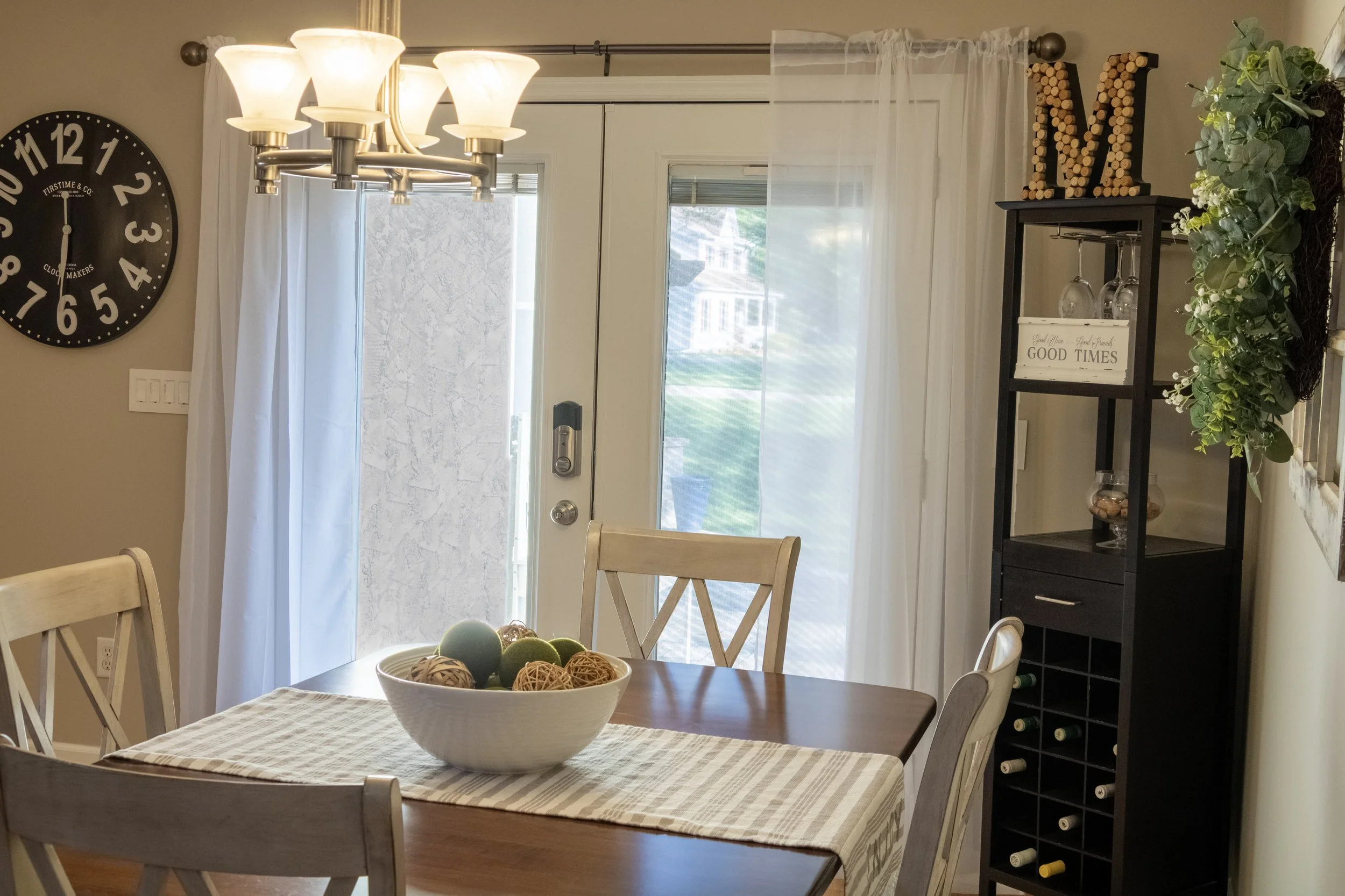 A dining area with a rectangular wooden table covered by a beige and white checked table runner, a white bowl filled with decorative balls, surrounded by light-colored wooden chairs with cross-back design. To the left, a large round wall clock with b
