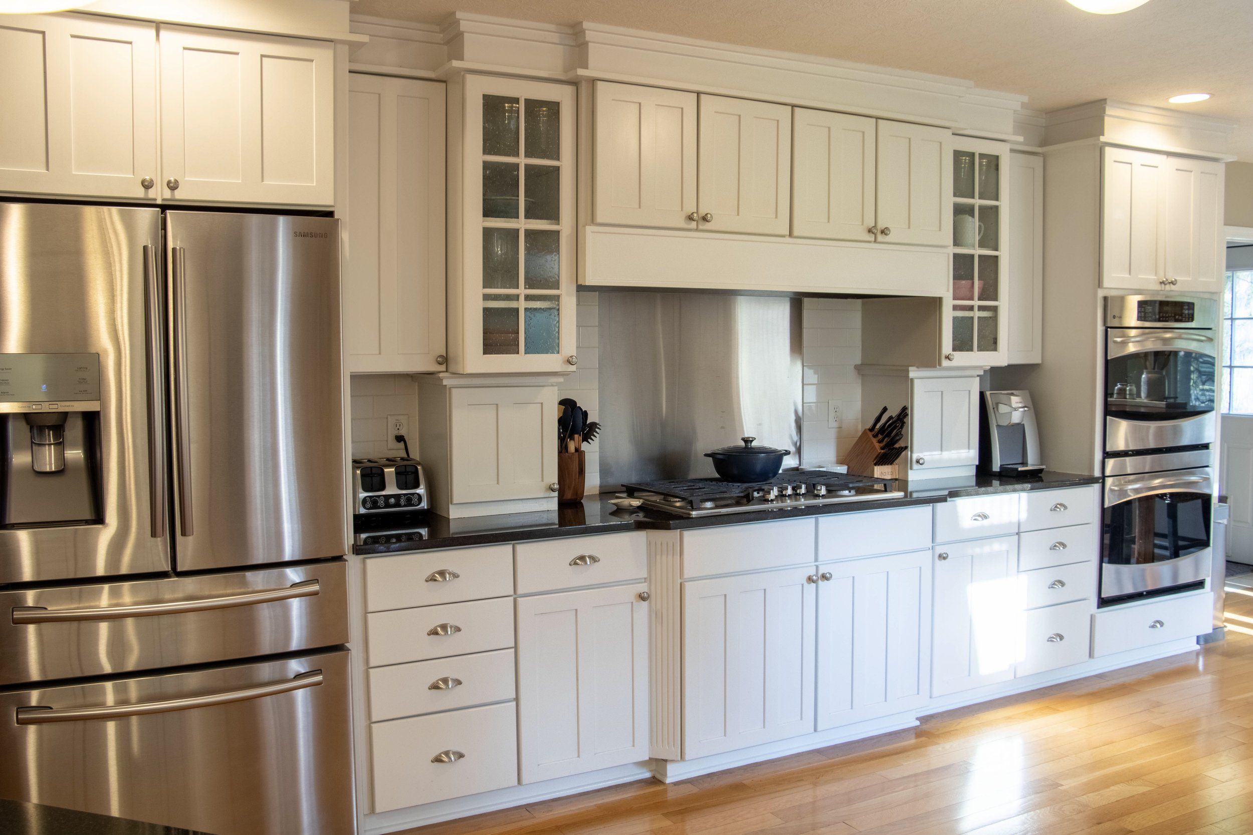 A kitchen with white cabinets, stainless steel refrigerator, oven, and microwave. The backsplash is white subway tile, and there is a window on the right side. The countertops are dark, and various kitchen utensils and a pot are visible.