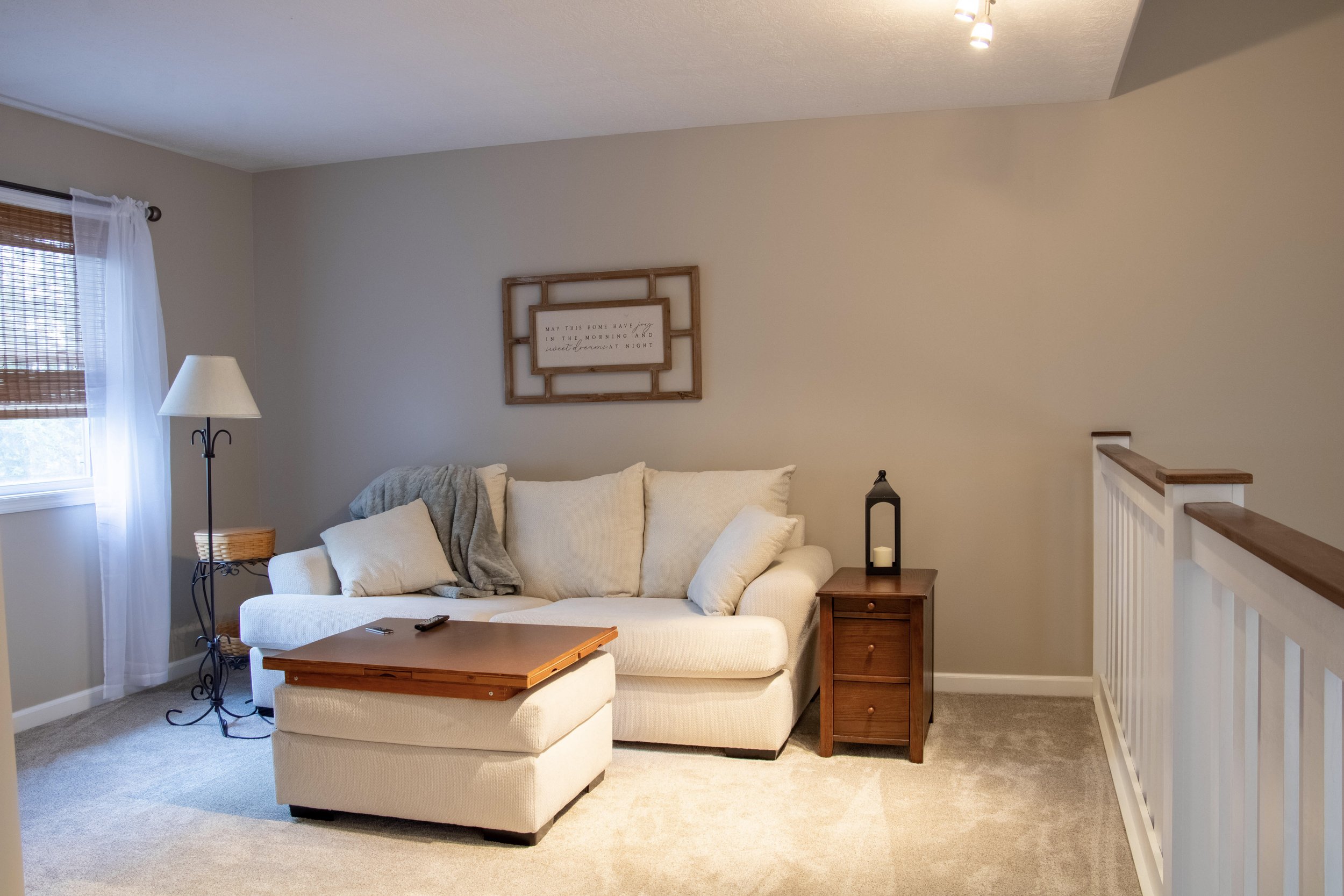 Living room with beige wall, white couch, wooden side table, floor lamp, wall art, window with blinds and sheer curtains, staircase railing.