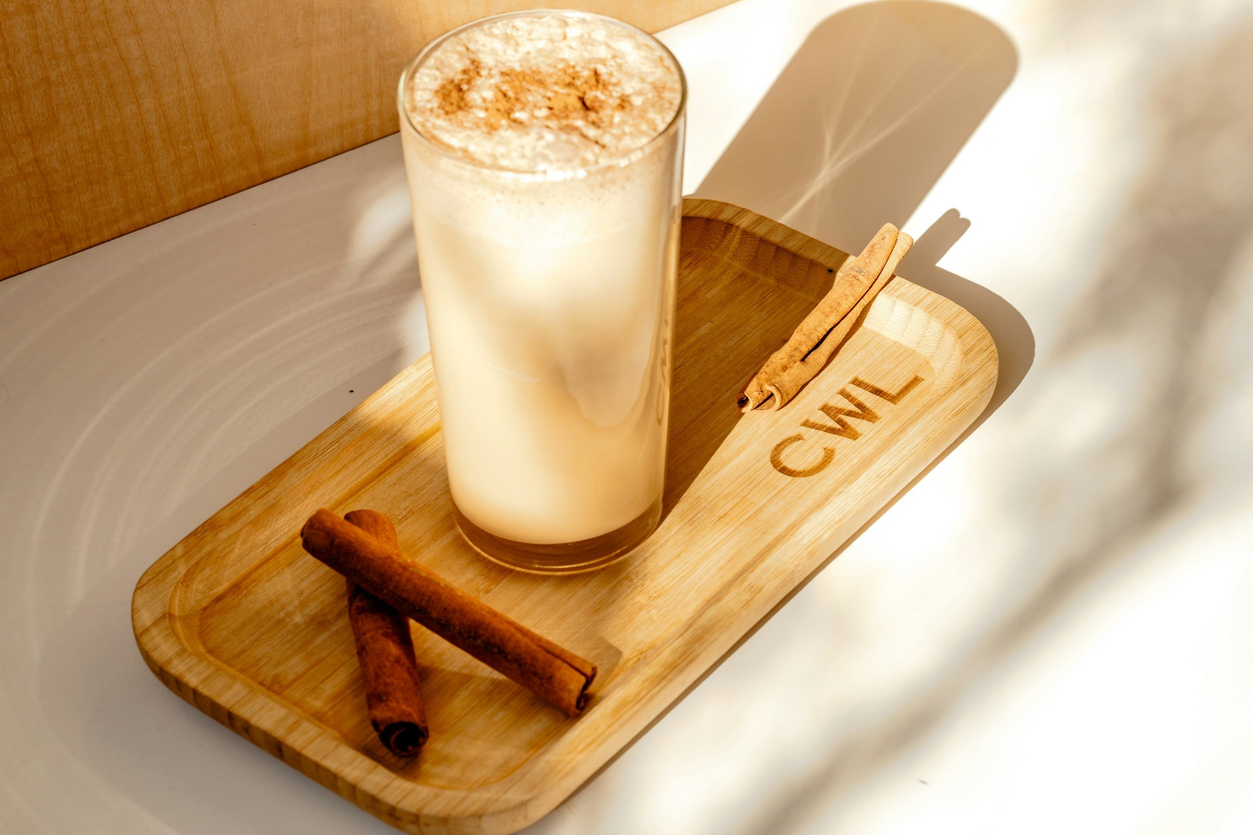 A tall glass of frothy vanilla latte topped with cinnamon sits on a wooden tray. Two cinnamon sticks and a star anise are also on the tray, which has the word 'CINNA' engraved on it. The tray is placed on a table with a white surface, with a wooden background behind.