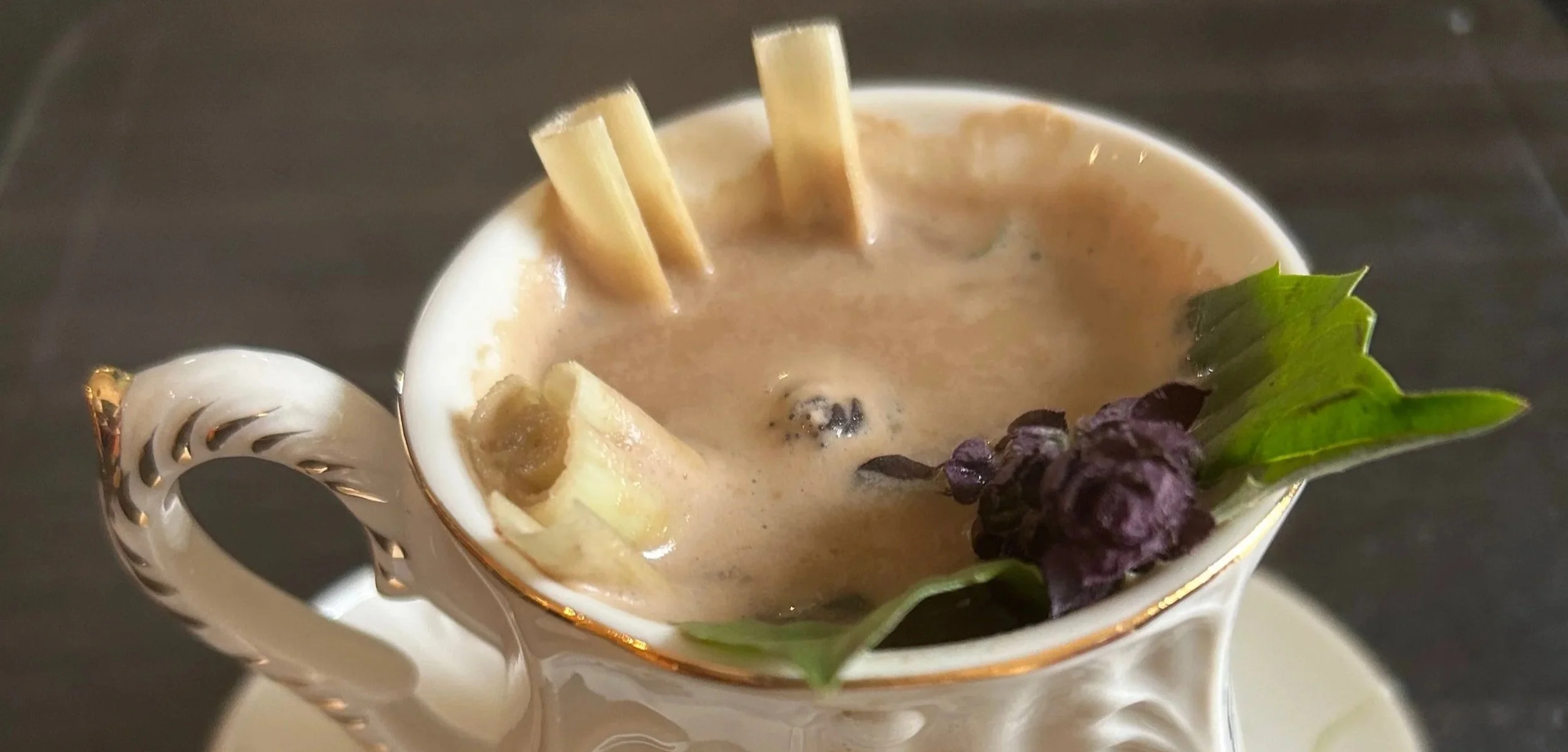 A cup of creamy soup garnished with a sprig of basil, black pepper, and pasta, set in an ornate white porcelain cup.