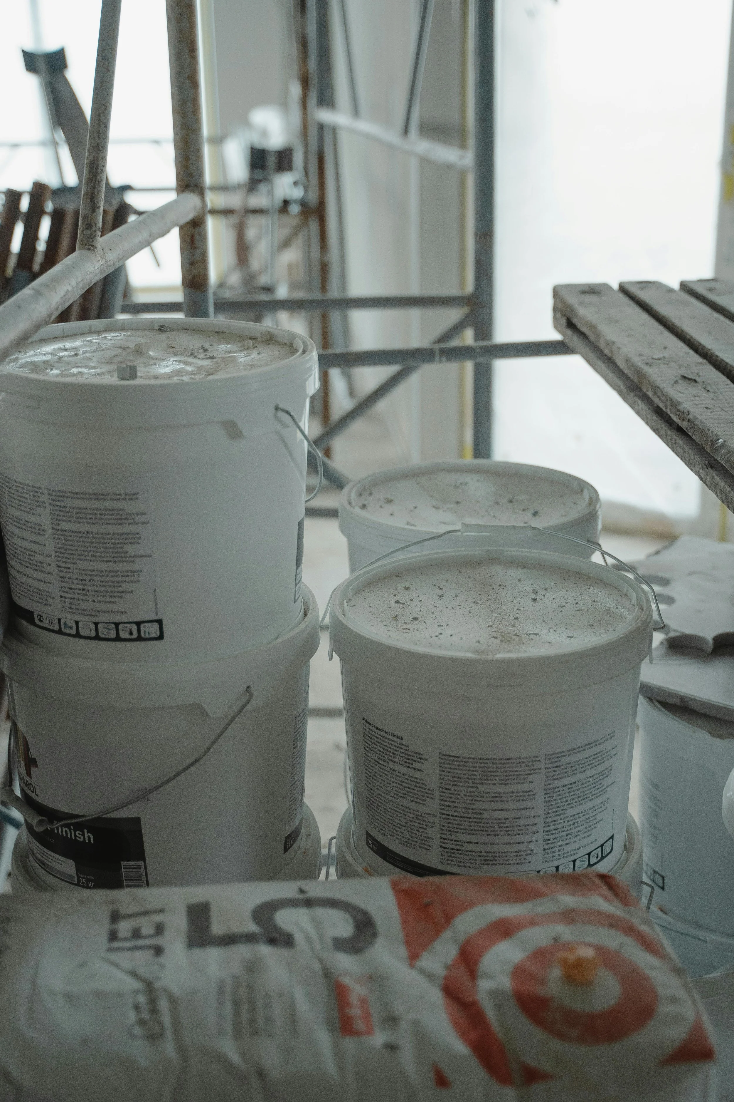 Multiple large white buckets with labels and a plastic bag with the Target logo, likely containing paint or construction material, in a room with scaffolding and construction supplies.