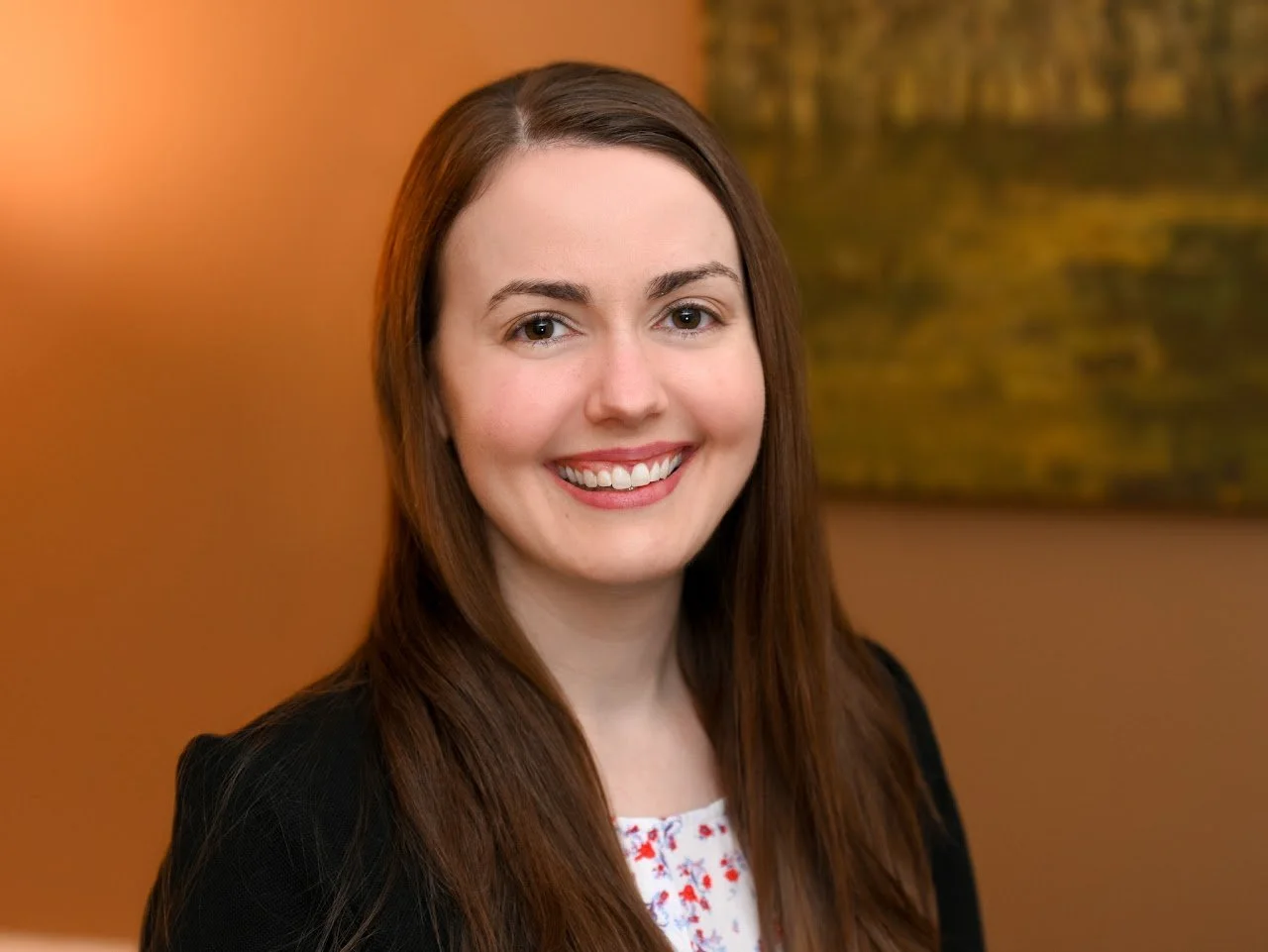 A woman with long brown hair, smiling, wearing a black blazer and a white blouse with red floral patterns, standing in front of an orange and green background.