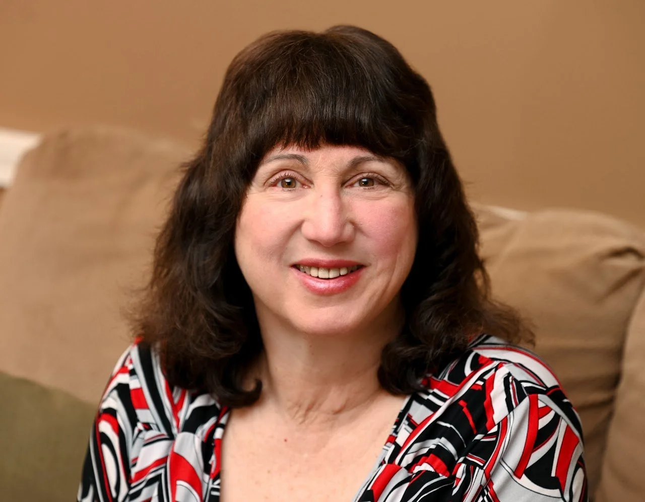 A smiling woman with medium-length dark hair and light skin, wearing a black, red, and white patterned top, sitting on a beige couch with a beige wall in the background.