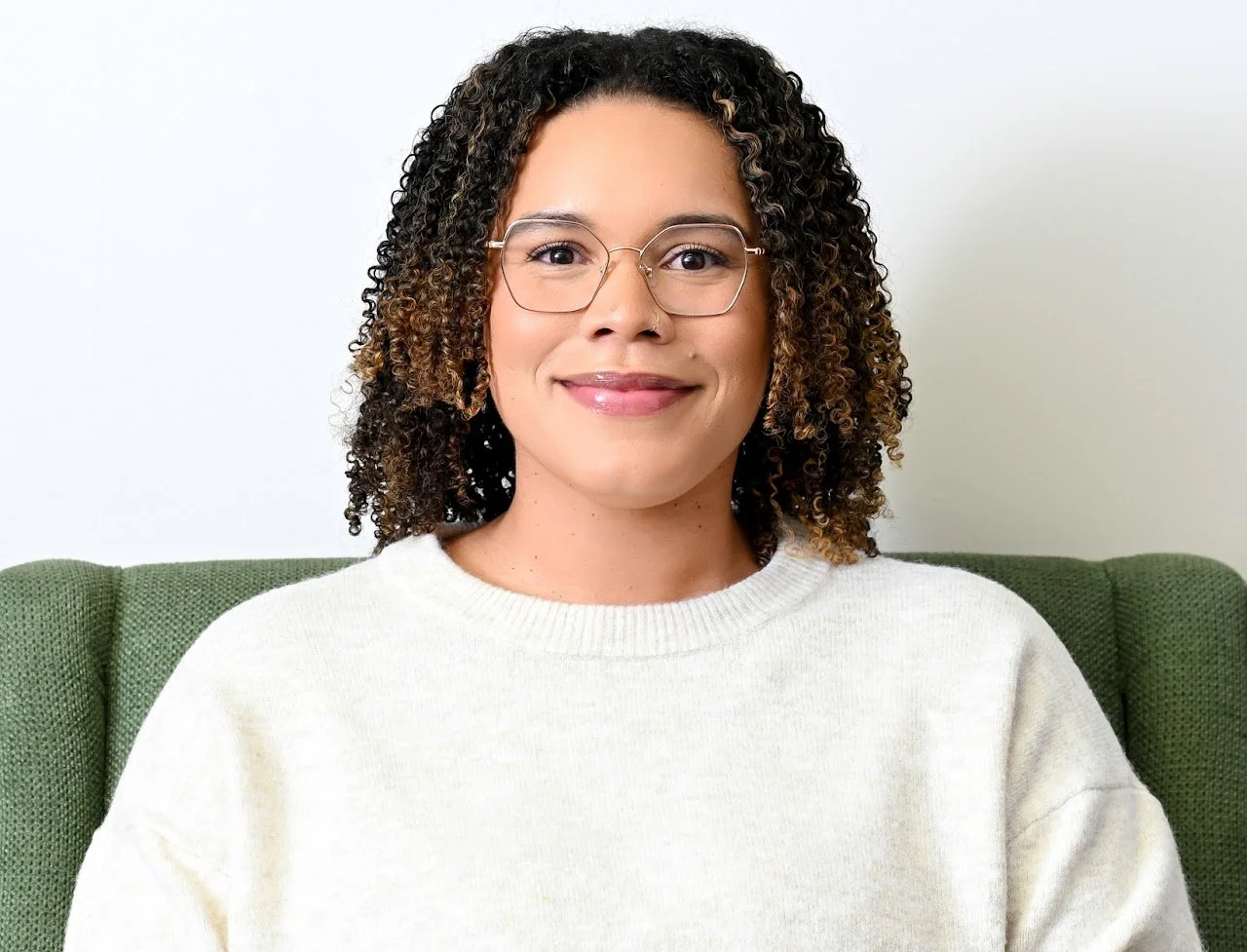 A woman with curly hair, glasses, and a light-colored sweater sitting on a green sofa against a plain white background.