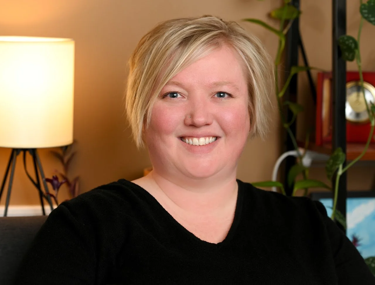 A woman with short blonde hair smiling, sitting in a room with a lamp and plants in the background.
